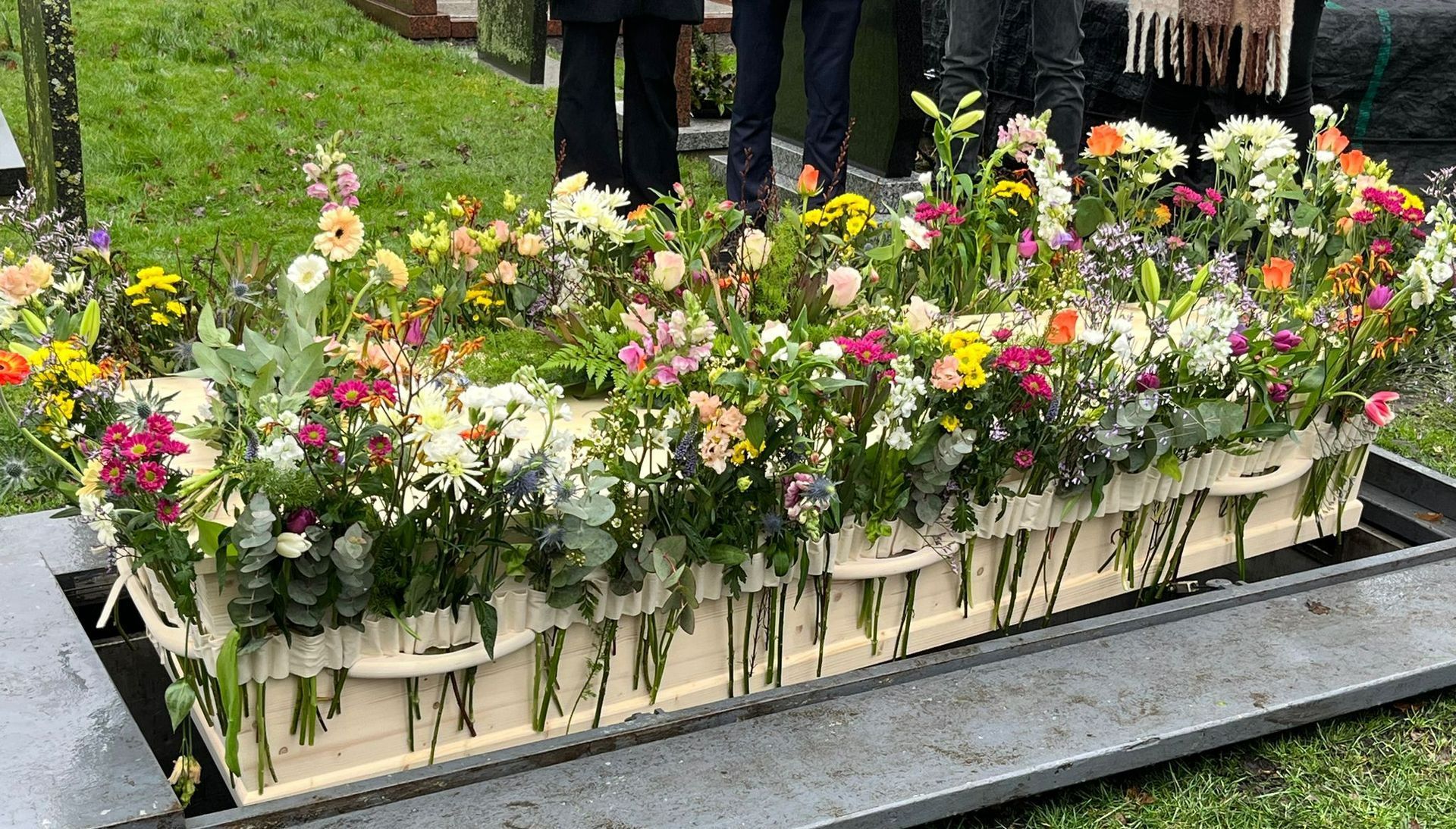 Een doodskist bedekt met bloemen staat op een graf. Blog de bloemenband.