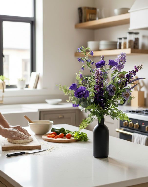 Keukenscène: Bloemen in een zwarte vaas op een wit aanrecht; iemand bereidt eten op een snijplank.