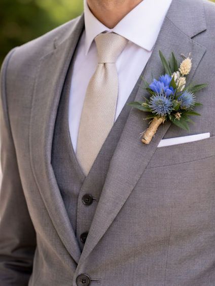 Man in grijs pak en vest met beige stropdas, wit overhemd en blauwe bloemencorsage.
