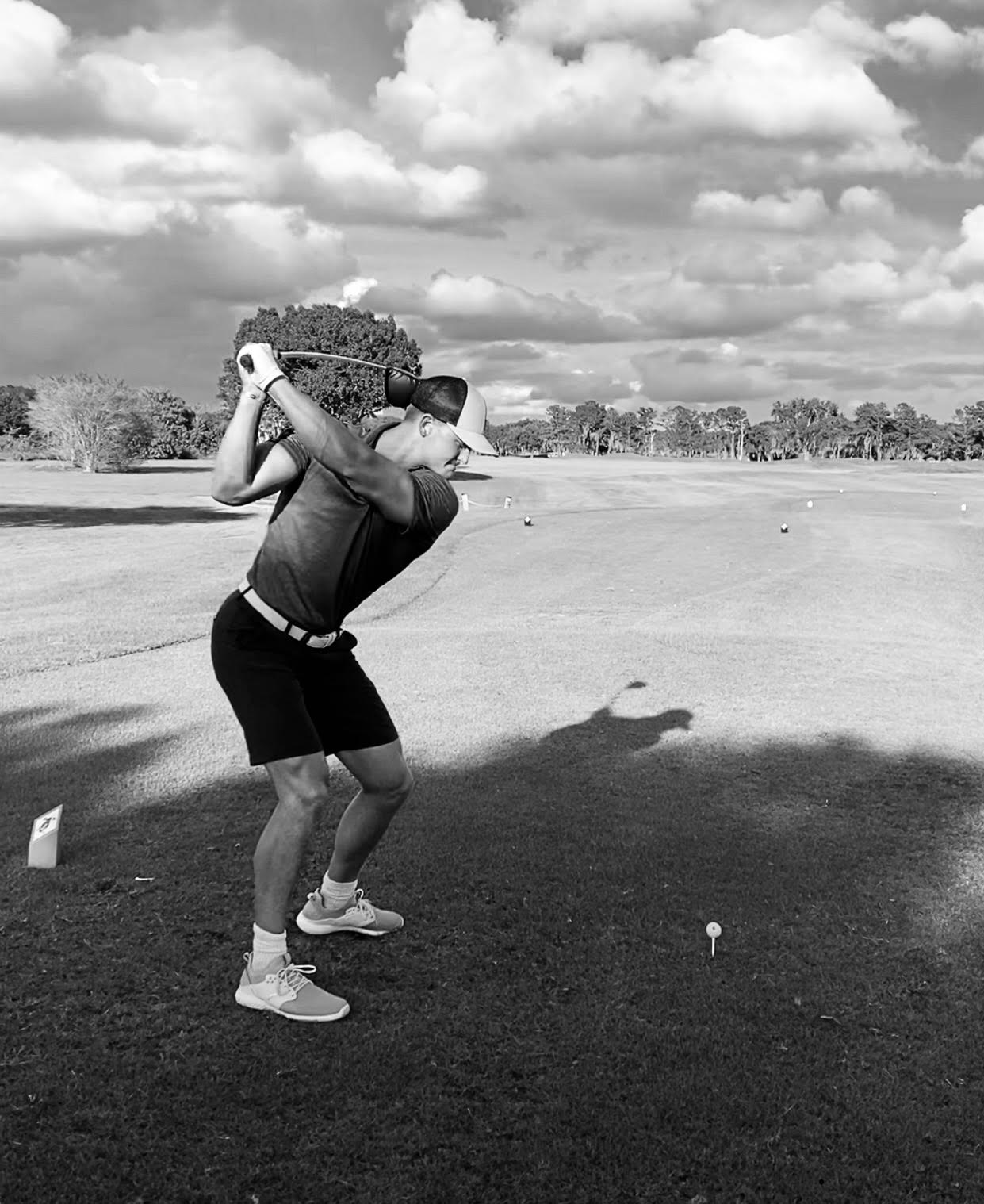 Golfer swings club at a driving range under a cloudy sky.