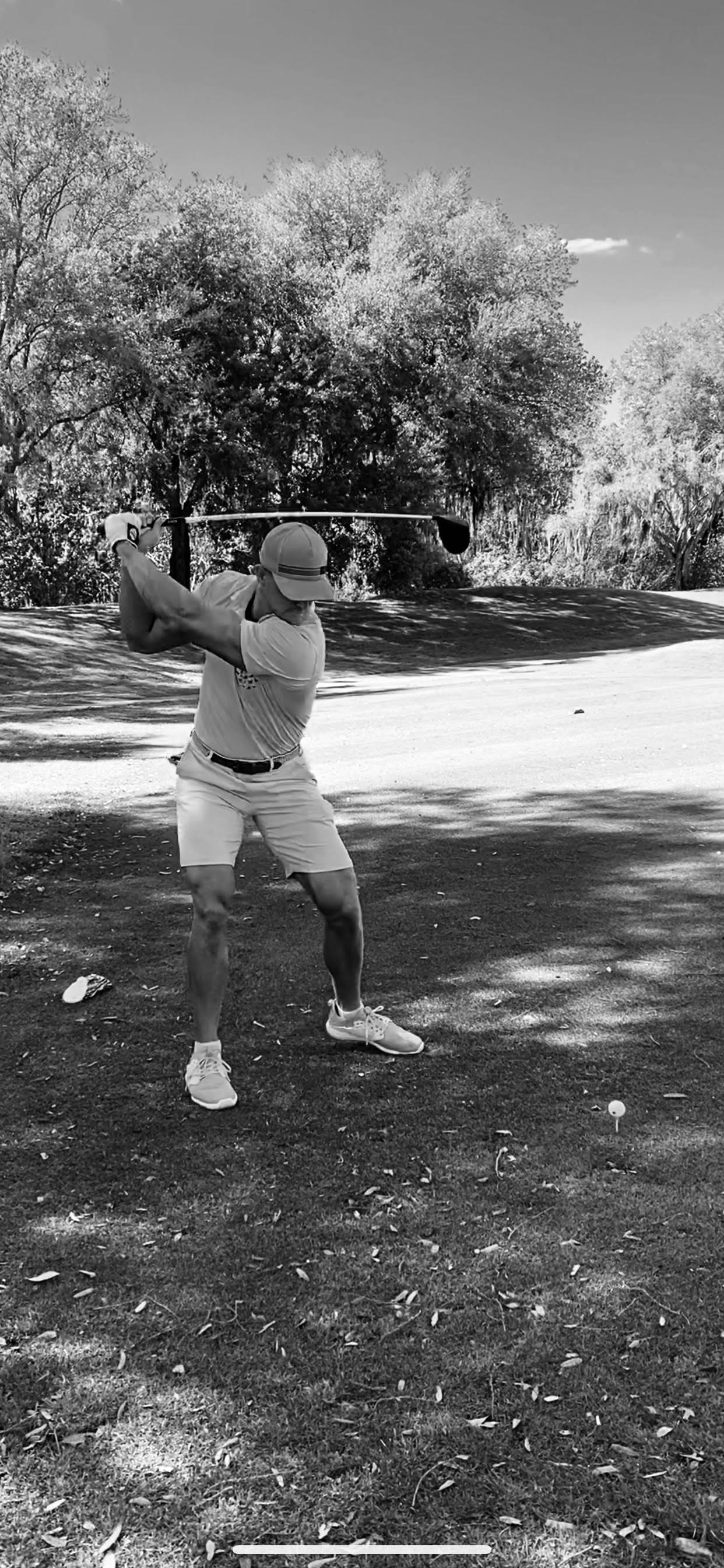 Man swinging a golf club outdoors under trees on a sunny day.
