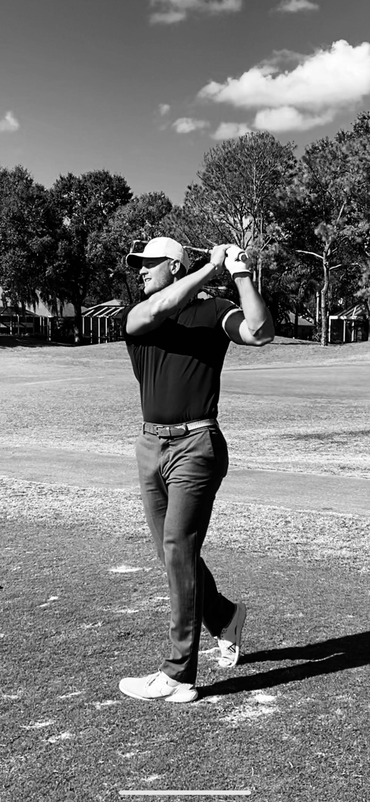 Golfer in mid-swing on a green golf course, wearing a cap, sunglasses, and dark polo shirt.