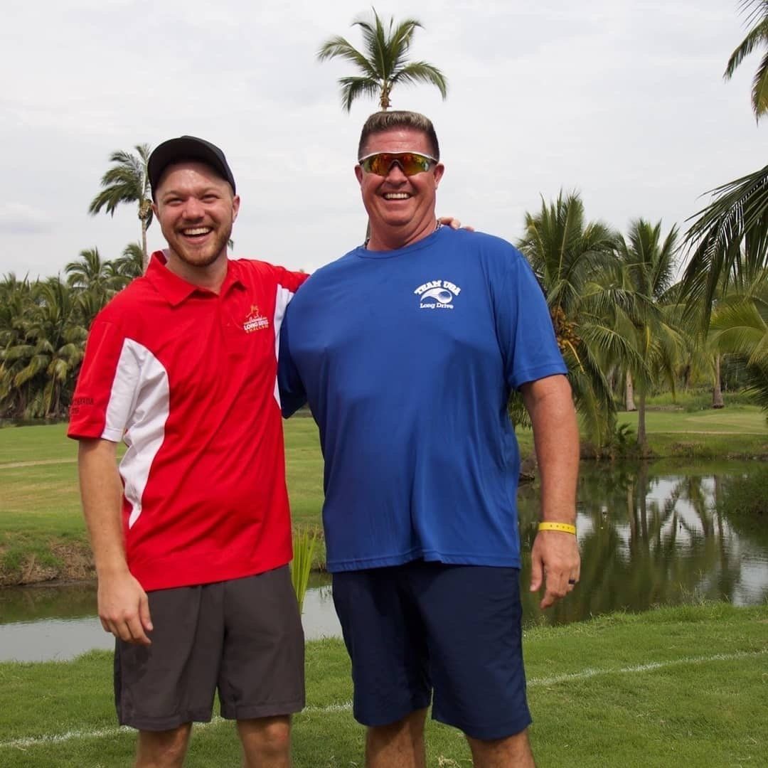 Two men smiling, arm-in-arm on a golf course with palm trees. One in a red shirt, one in blue.