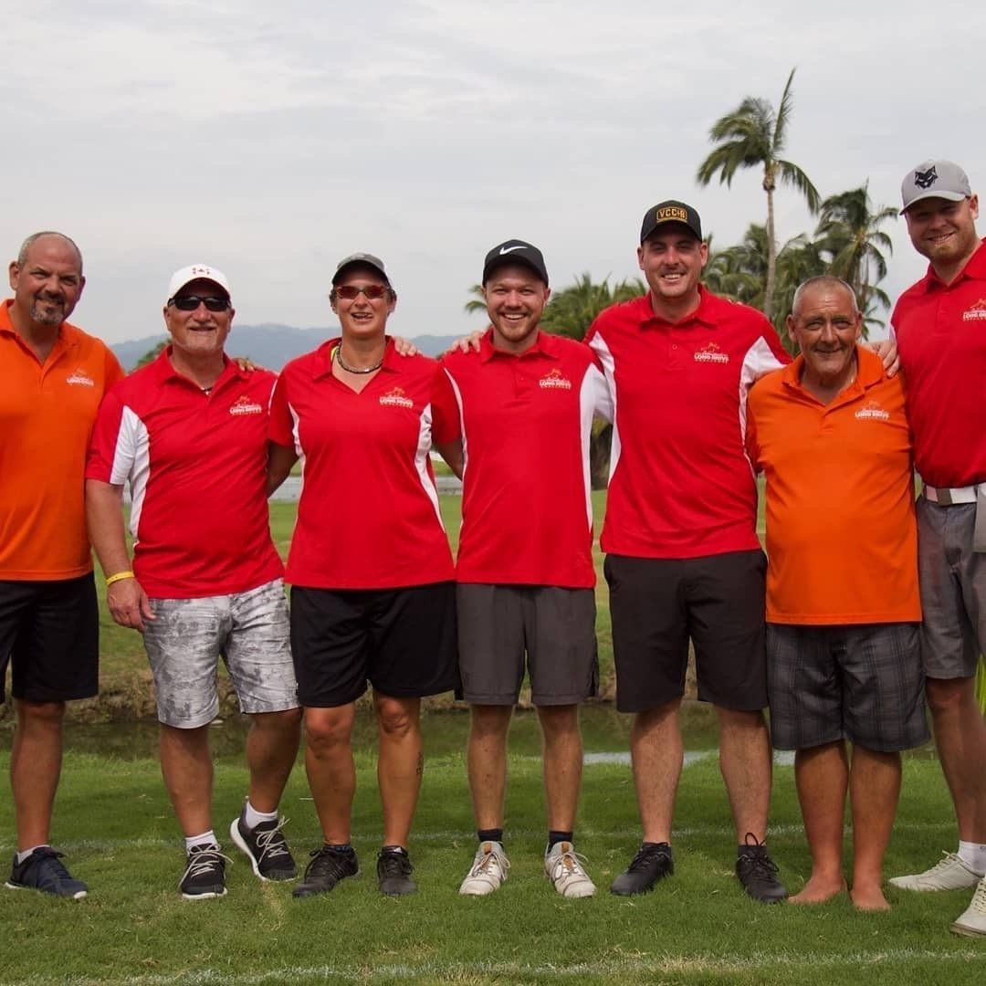 Group of seven people wearing red or orange shirts, posing outdoors with arms around each other.