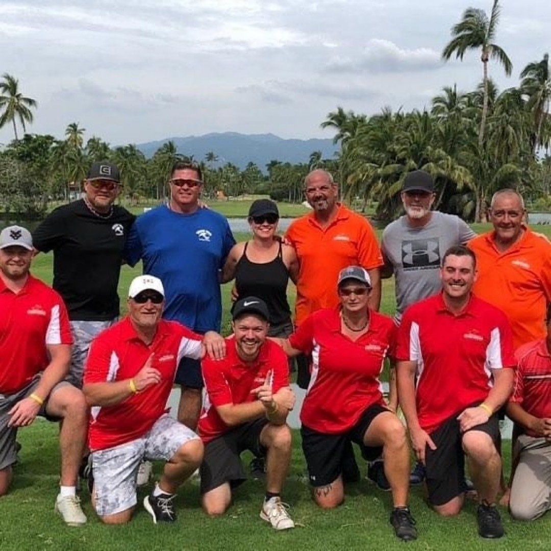 Group of people posing on a golf course; many wearing red shirts; tropical setting with palm trees and mountains.