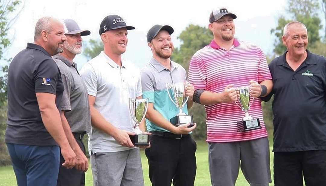 Group of men holding trophies, smiling, outdoors. Some wear golf attire. Green background.