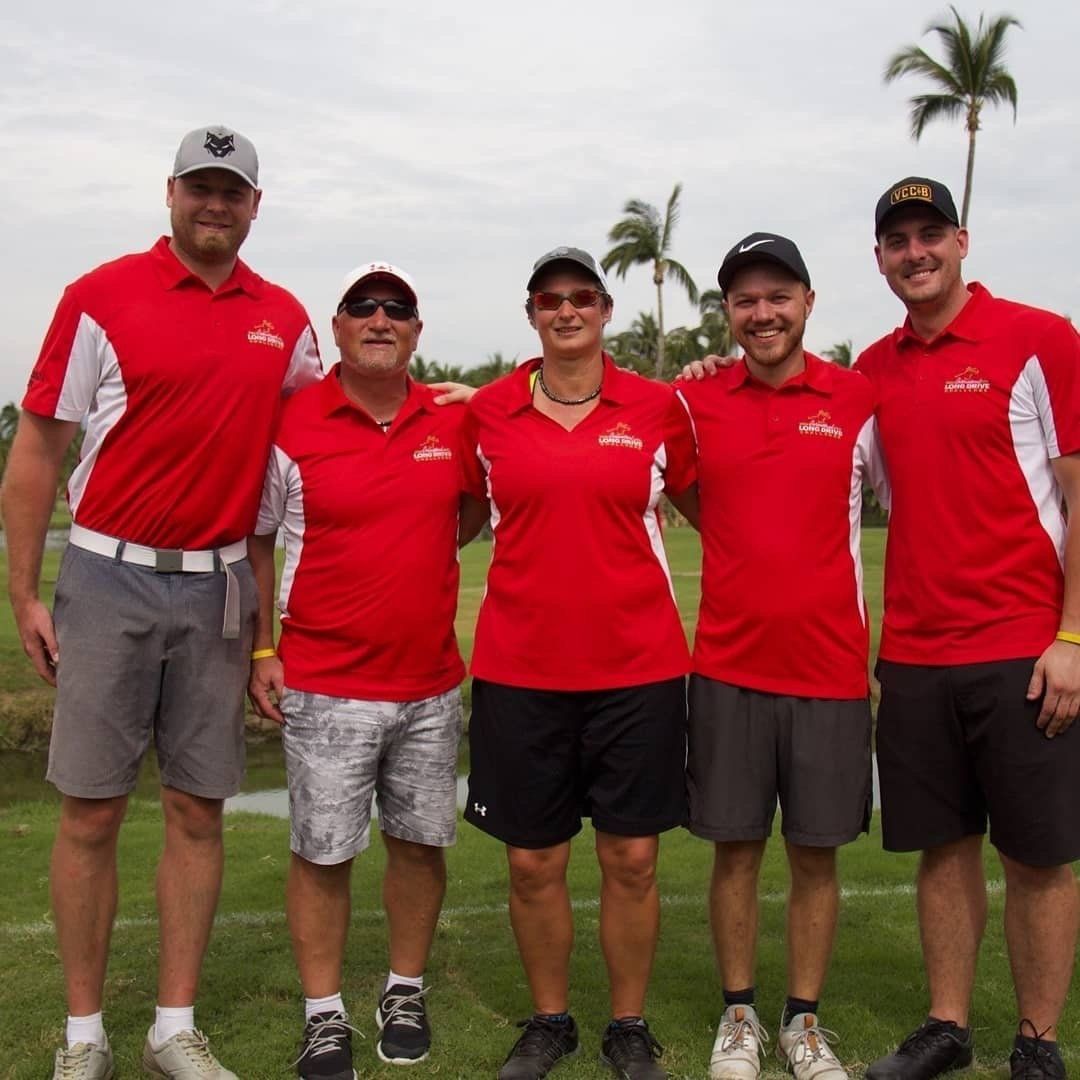 Five people wearing red shirts posing on a green field. Palm trees in the background.