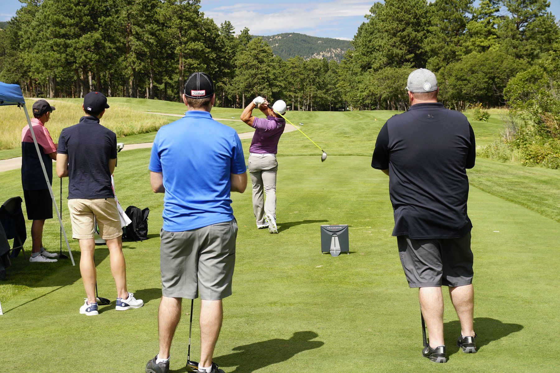 A group of men are standing on a golf course watching a man swing a golf club.