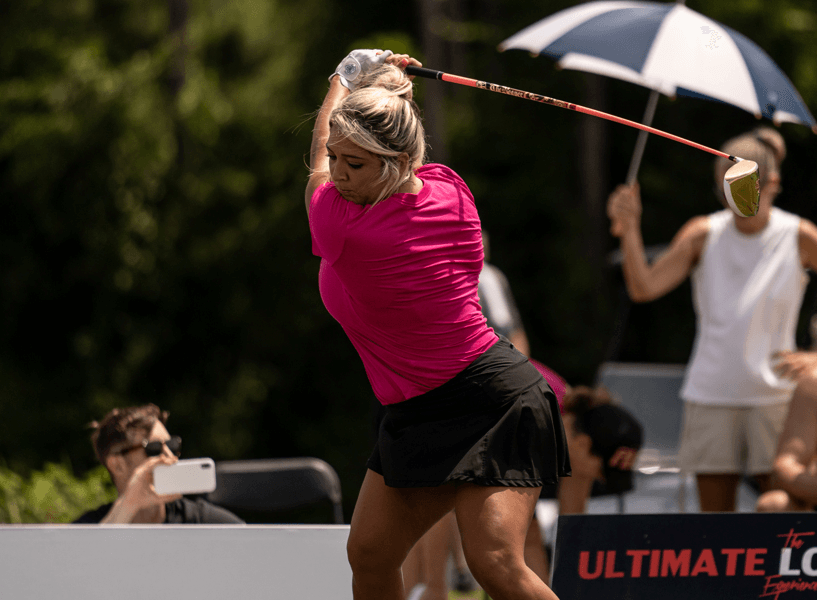 A woman in a pink shirt and black skirt is swinging a golf club.