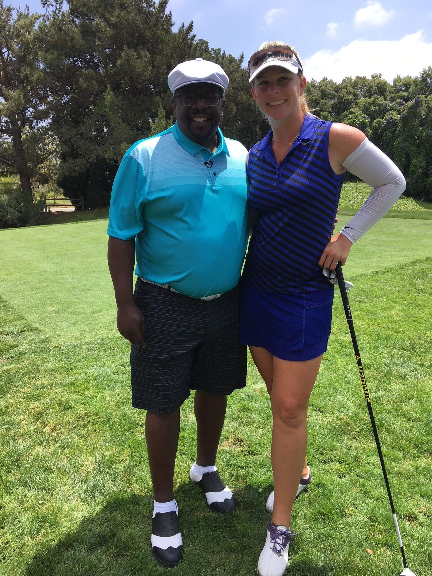 Two people pose on a golf course. Man in turquoise shirt, woman in blue outfit, holding a golf club.