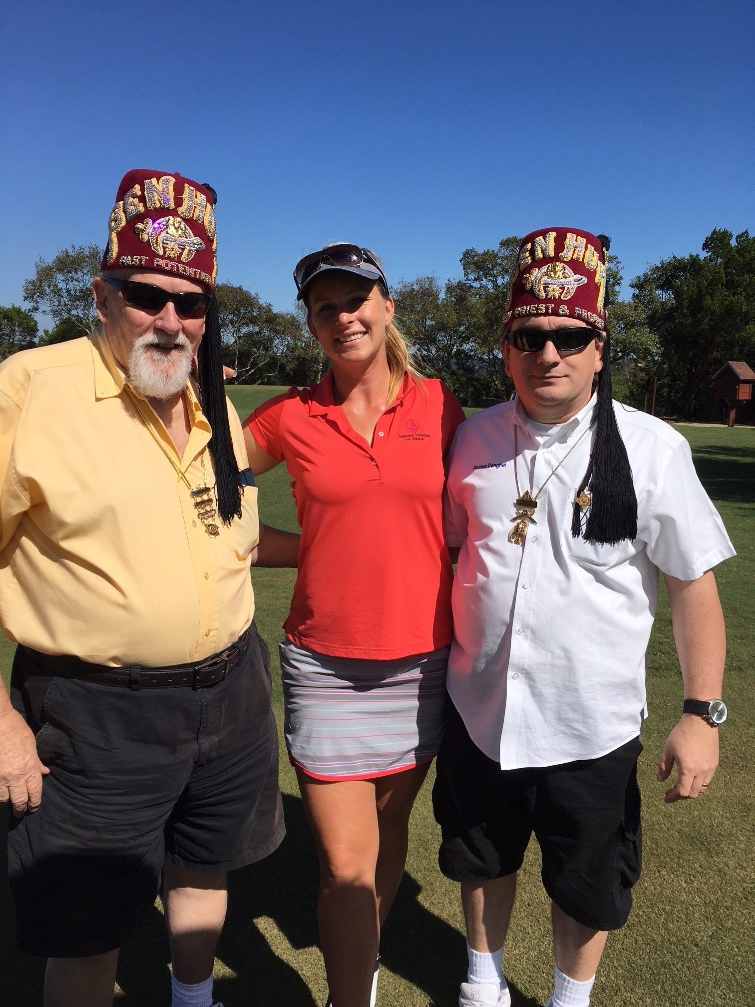 Three people on a golf course: two men in fezzes and sunglasses flank a woman in a red shirt and visor. Sunny day.