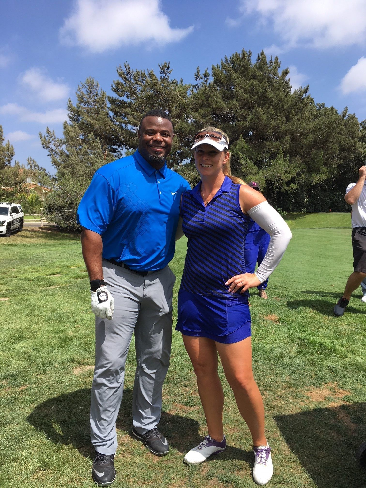 Man and woman posing on a golf course. Man in blue shirt, grey pants. Woman in blue golf attire, visor. Sunny day.