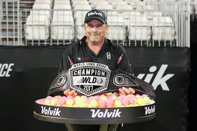 Man holding a champion belt, standing in front of a table with golf balls, Volvik sign behind him.