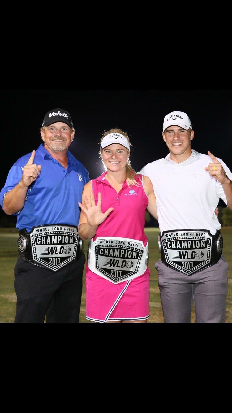 Three people wearing championship belts, outdoors at night. Woman in pink points five fingers.