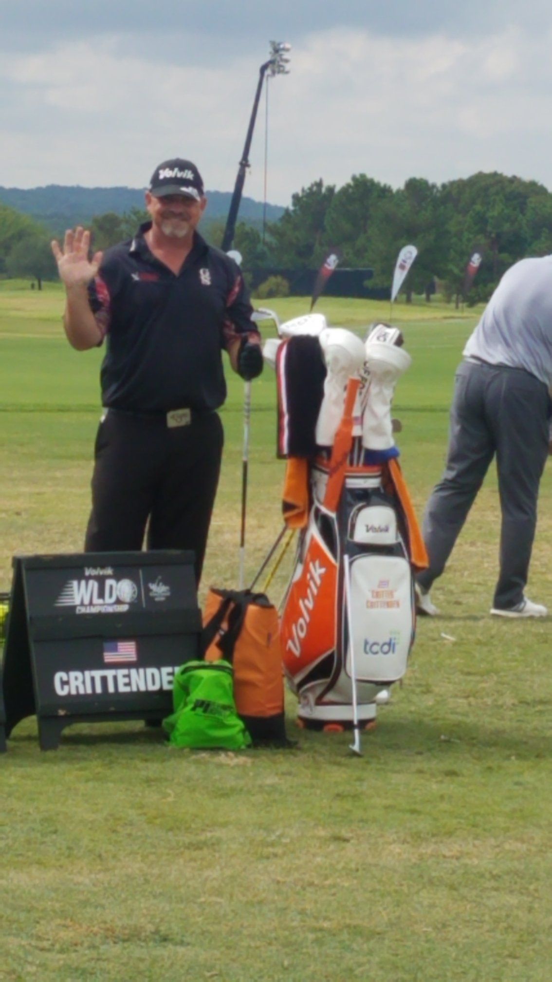 Golfer waves, standing by his golf bag and a sign at a golf course.