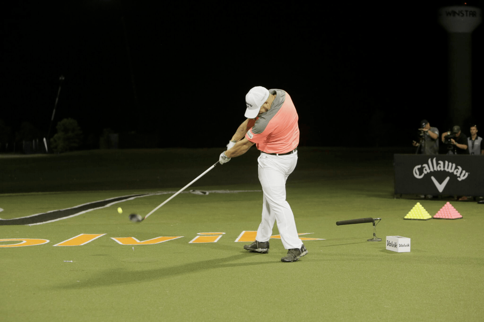 Golfer swinging a club at night, hitting a golf ball. White hat, orange shirt, white pants.