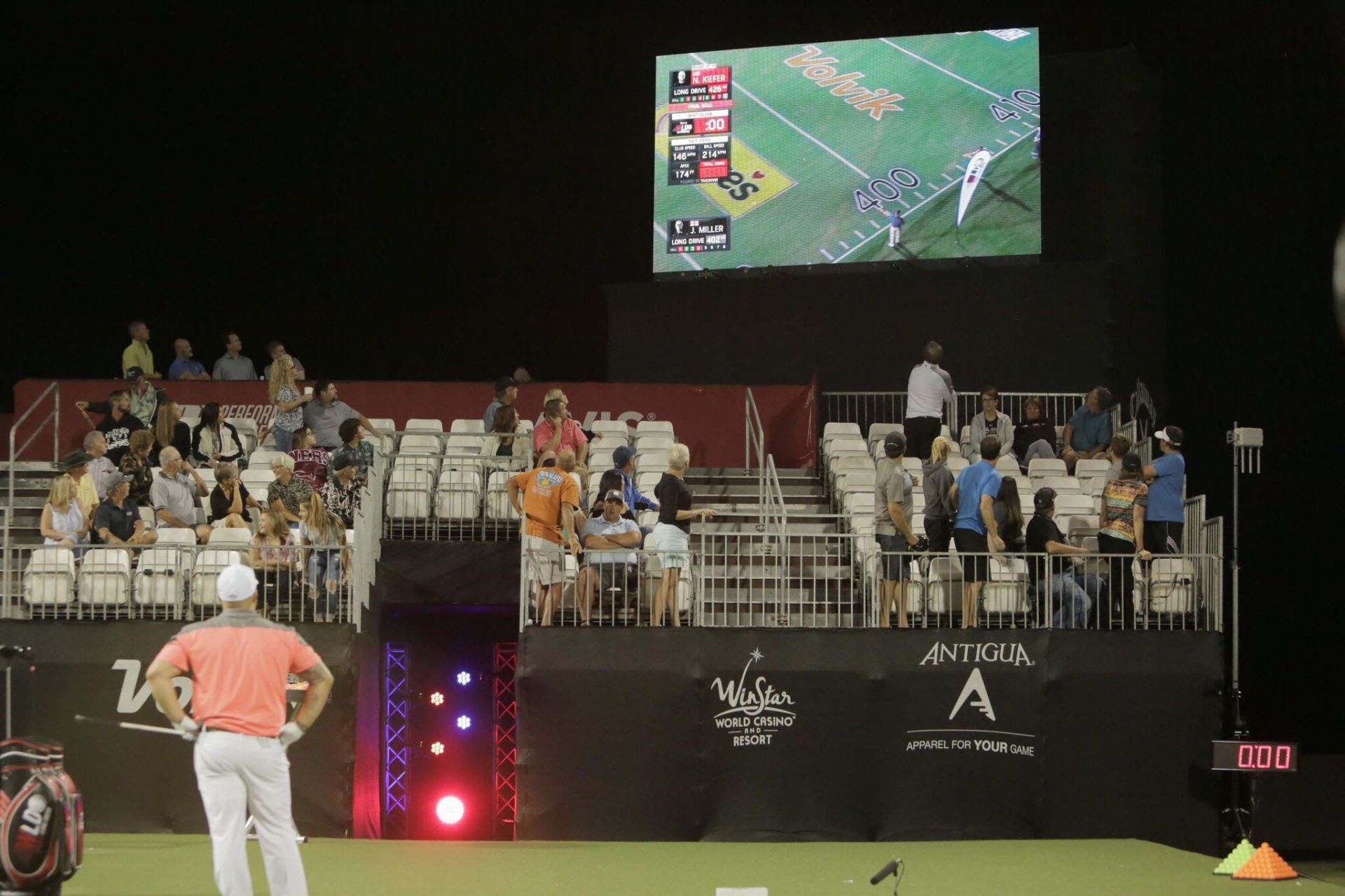 Golfer on green, facing audience in bleachers, watching a screen showing a golf course.