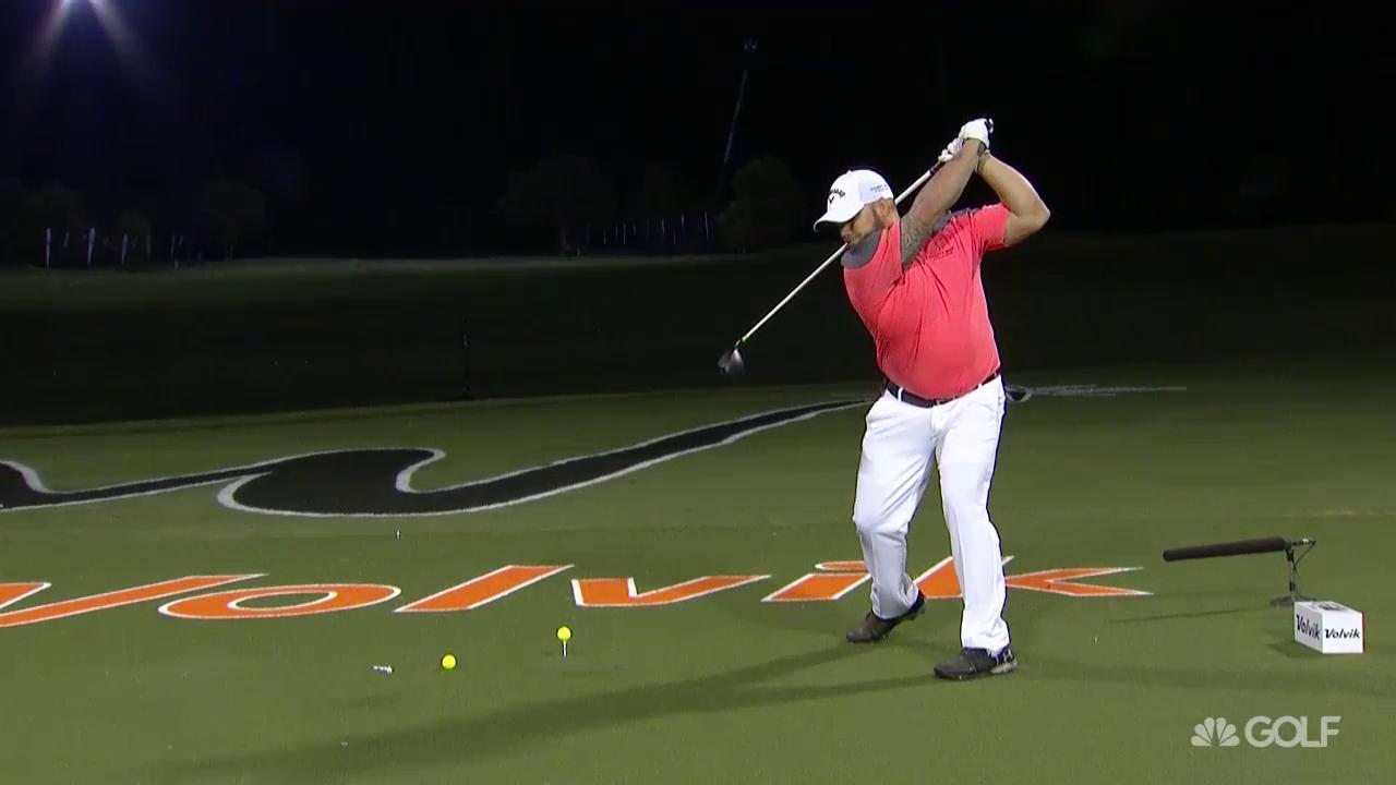 Golfer in red shirt swings golf club at night, aiming toward the right on a green field.