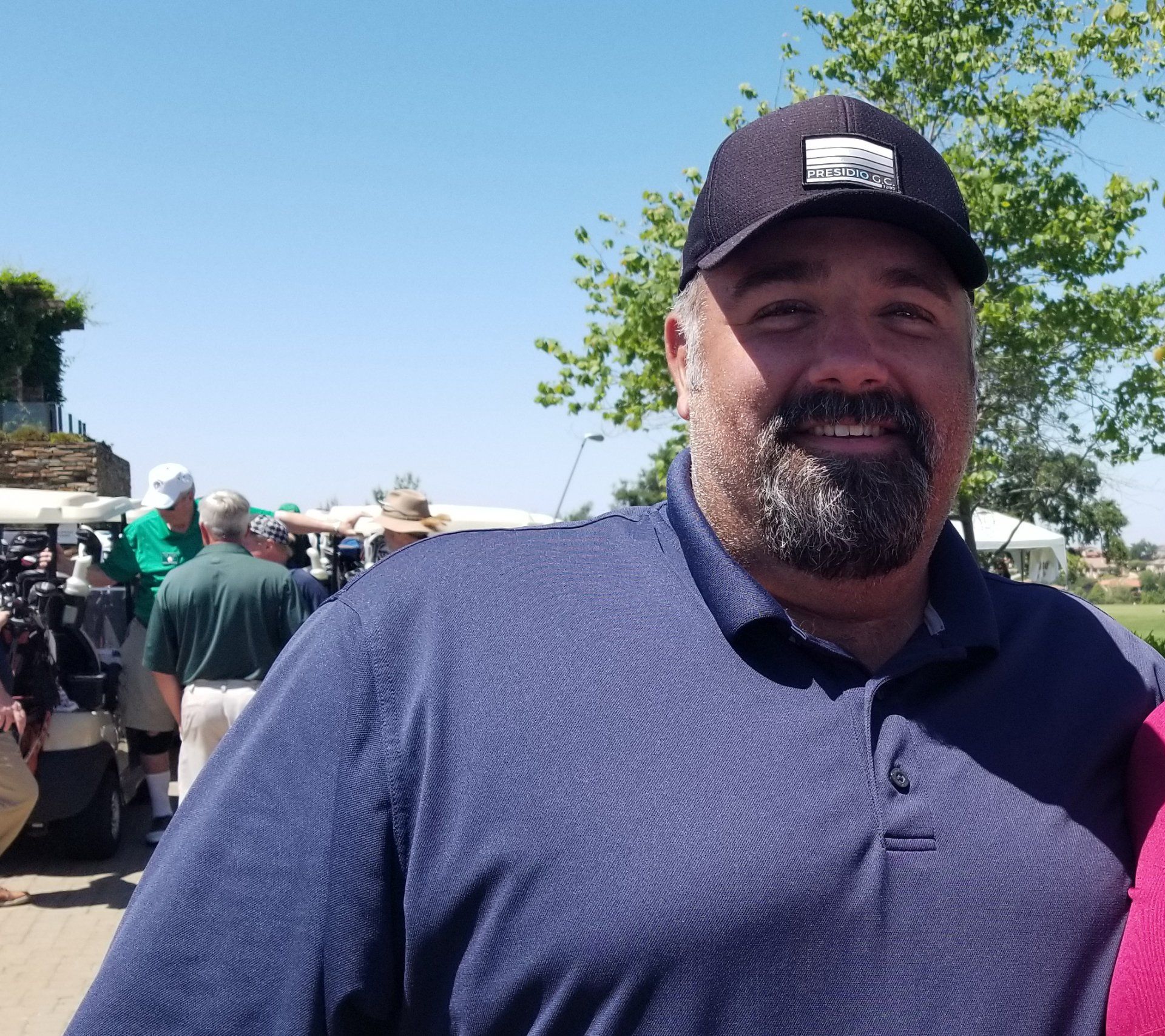 Man wearing a dark baseball cap and navy polo shirt smiles outdoors, with a golf course in the background.