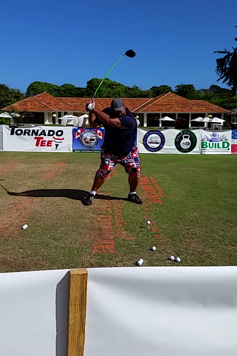 Golfer swinging a club on a driving range, wearing a cap, floral shorts, with logos in the background.