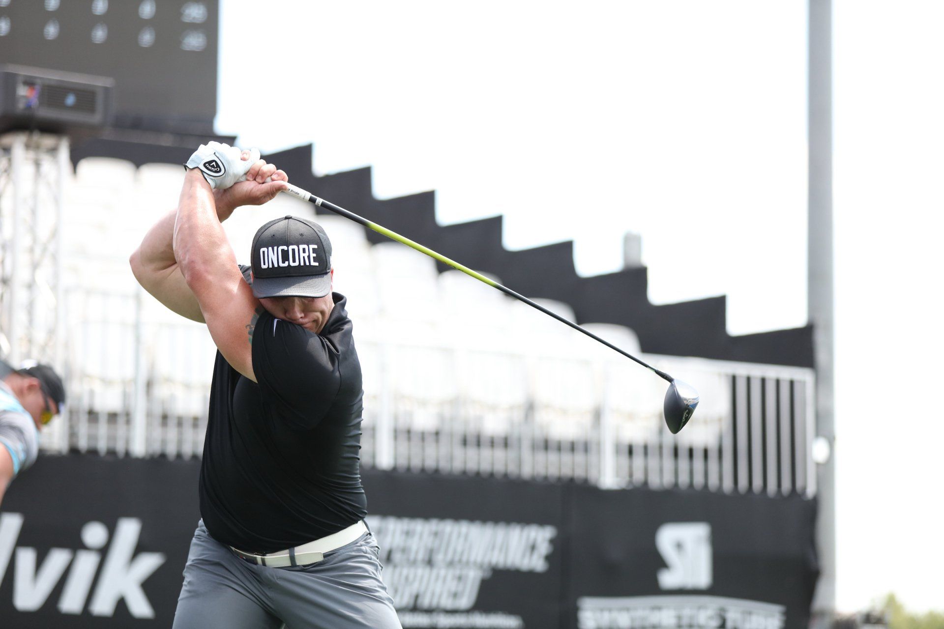 Golfer mid-swing on a course; black shirt, cap, grey pants; blurred background with scoreboards and structure.