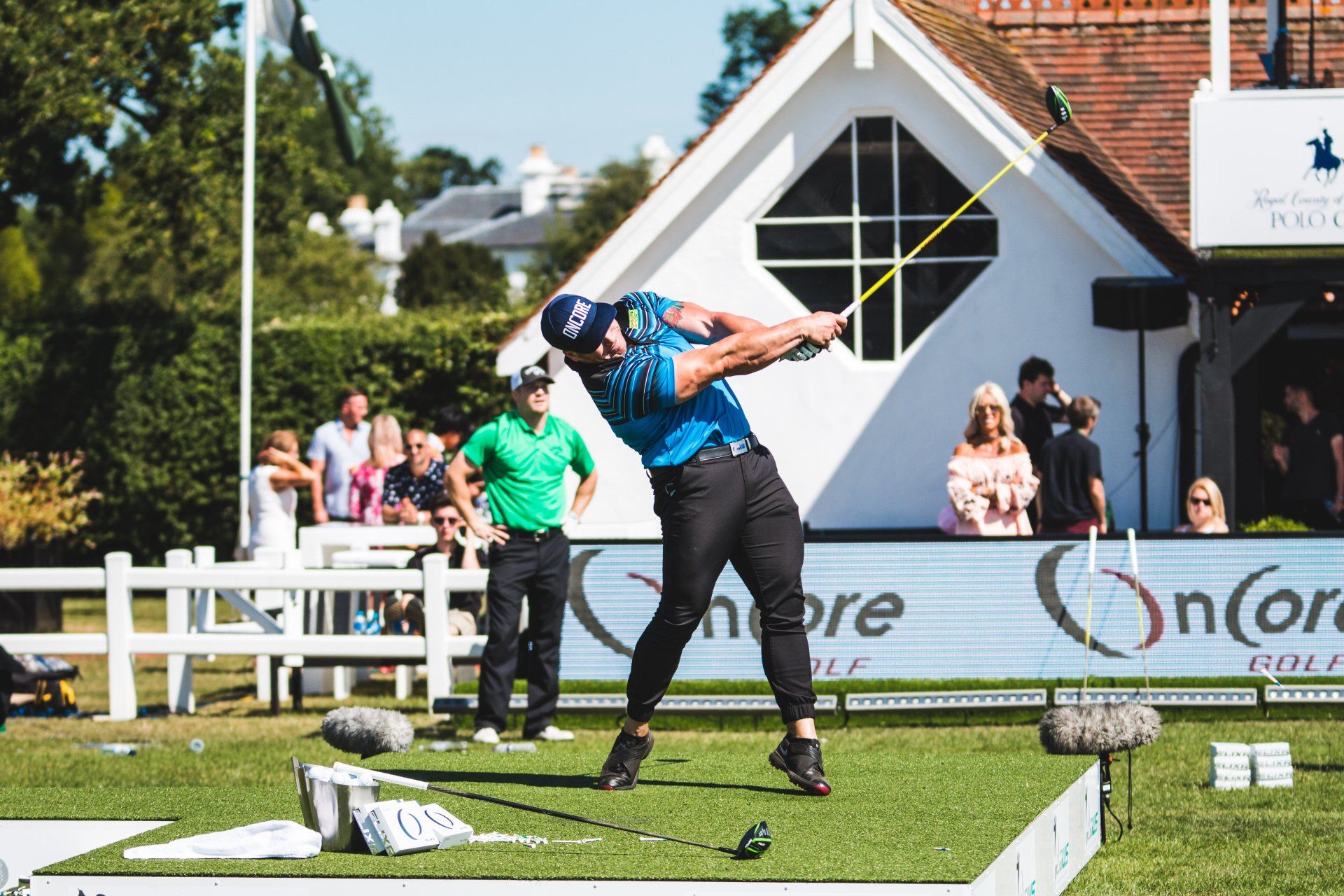Golfer swinging club on a tee box, near a white building with spectators.