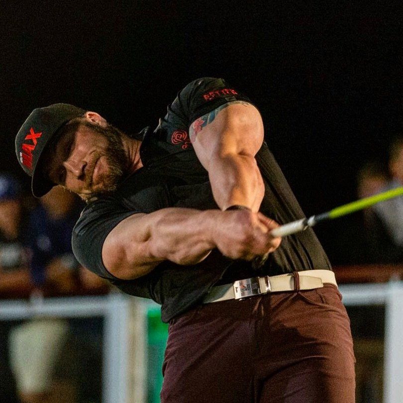 Man swinging a golf club, with large arm muscles, wearing a black cap and shirt, brown pants.