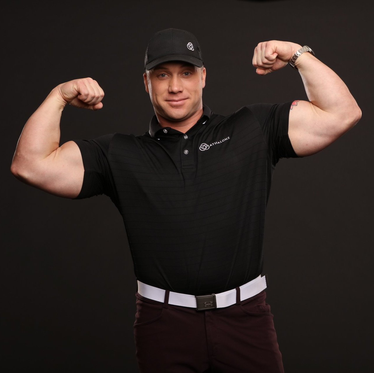 Man flexing large biceps, wearing a black shirt, cap, and white belt.