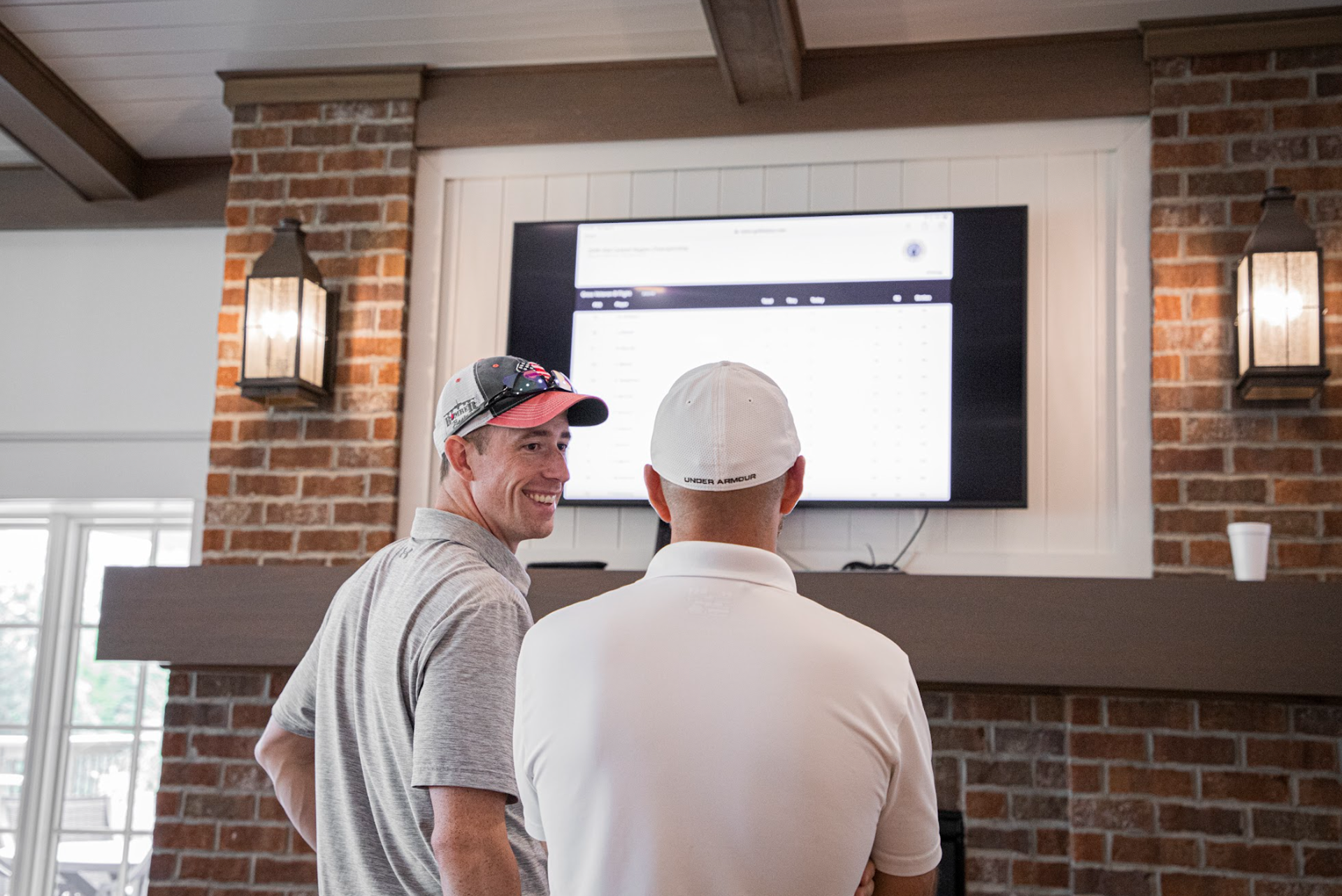 Two golfers view a live leaderboard in the clubhouse at a charity golf tournament.