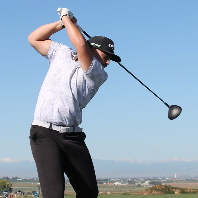 Man wearing black hat swinging a golf club, background: golf course.