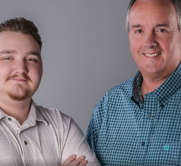 Two men smiling at the camera, one young with a goatee, the other older in a blue patterned shirt.