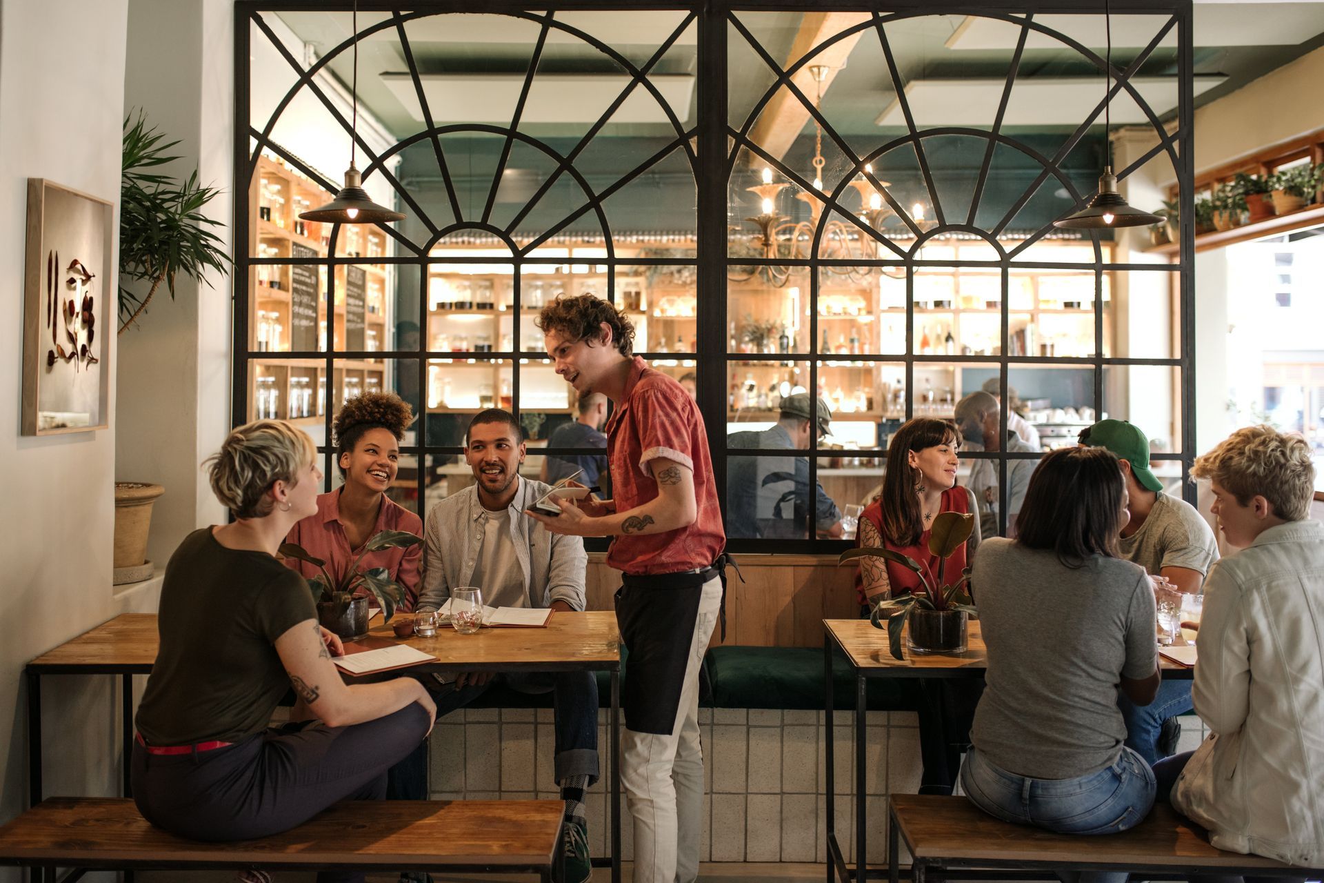 Customers seated at tables in a cafe, server taking order. Black window frames, neutral tones.