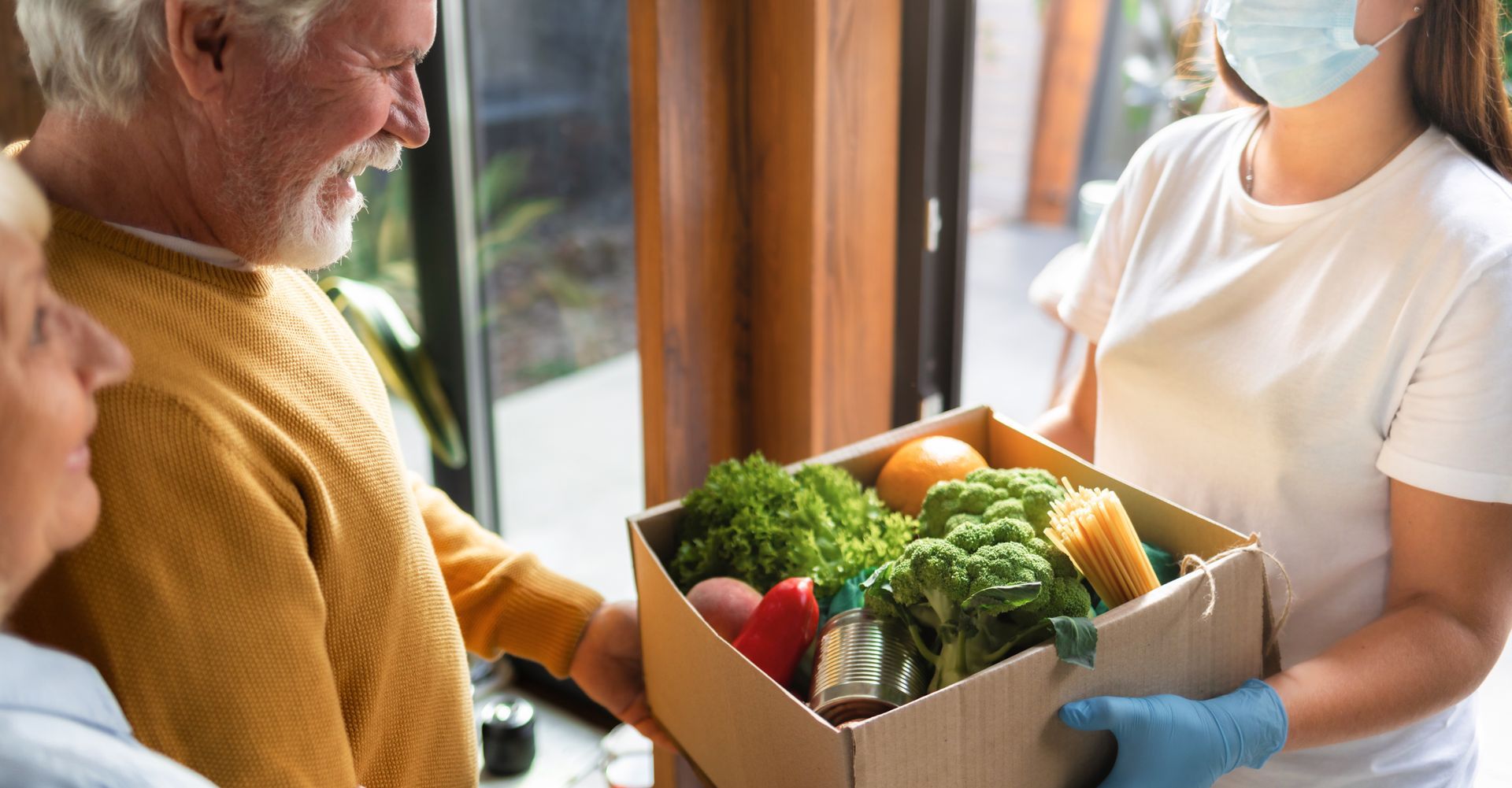 Person delivering a box of groceries to an elderly couple at their doorway.