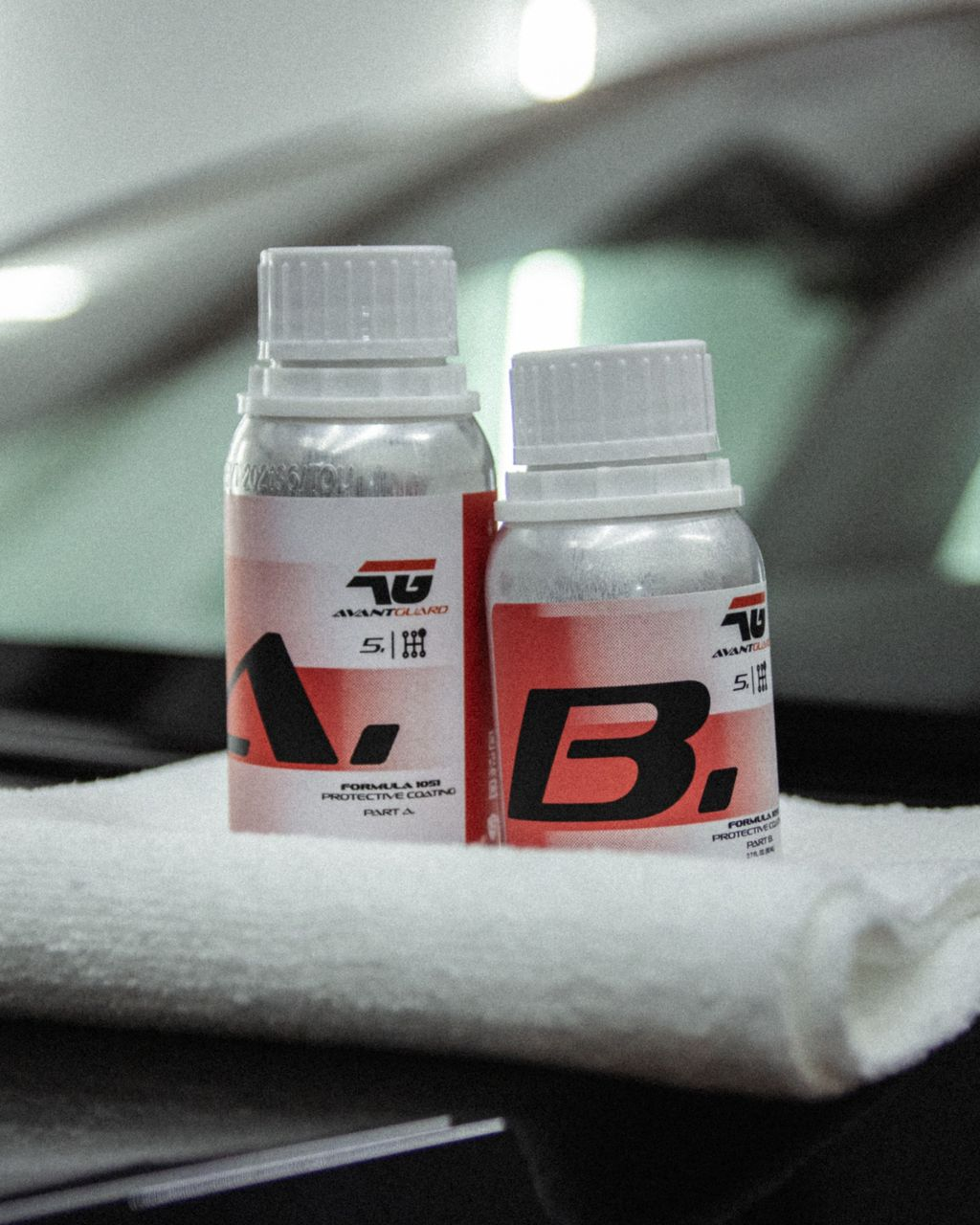 Two small red-and-white bottles labeled A and B on a white towel, with a car in the background