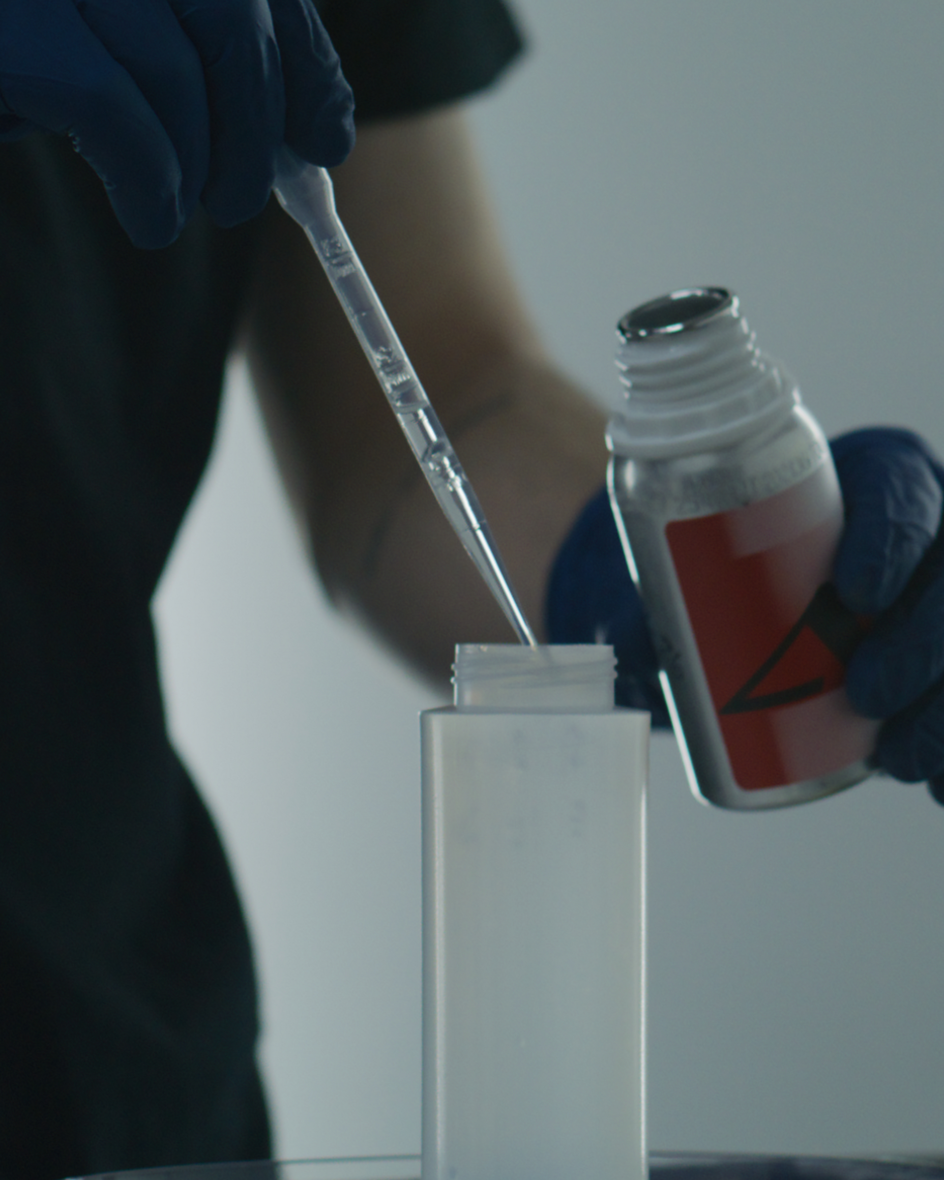 Gloved hands using a pipette and bottle to fill a white container in a lab setting
