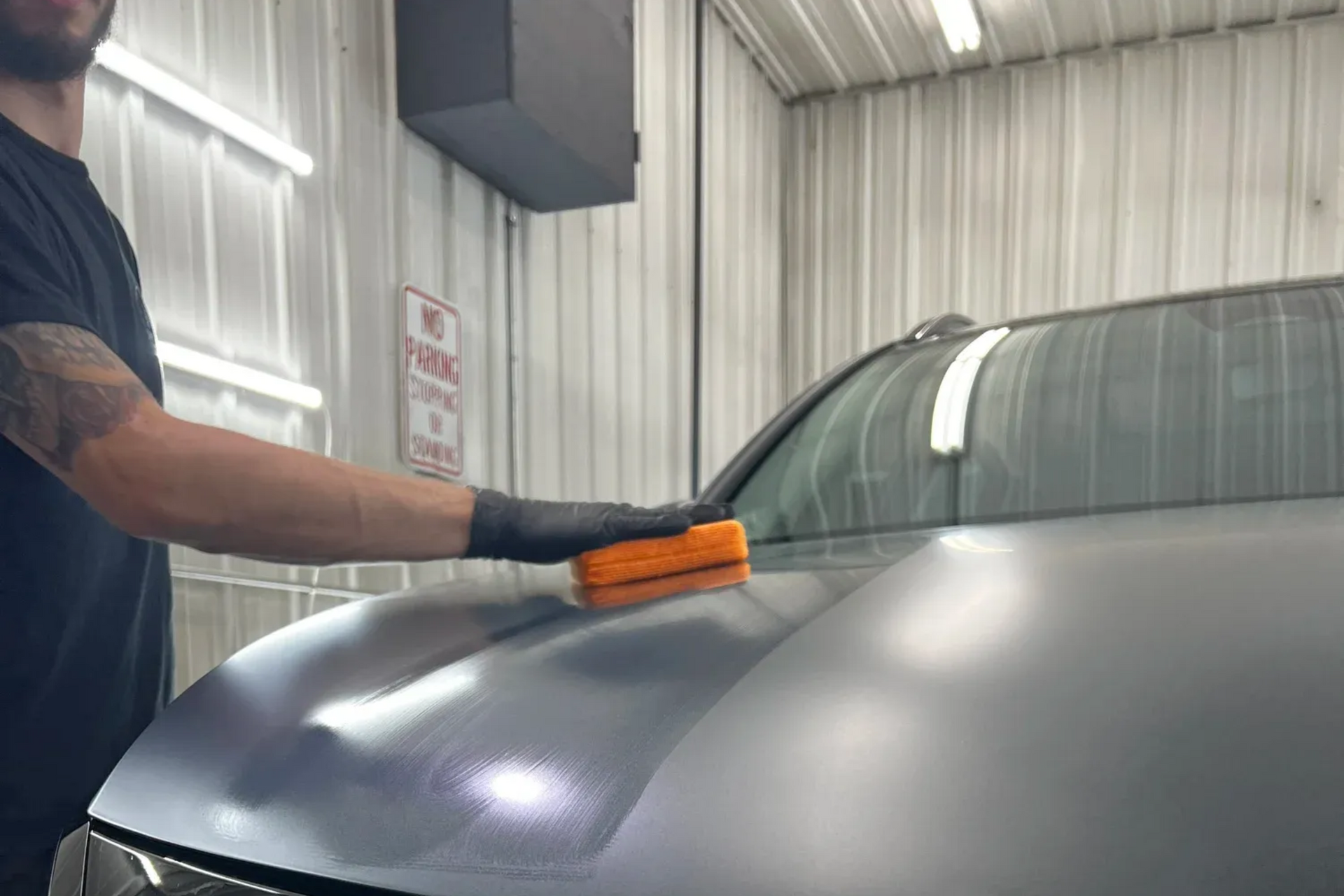 Person polishing a gray car hood with an orange applicator in a garage.