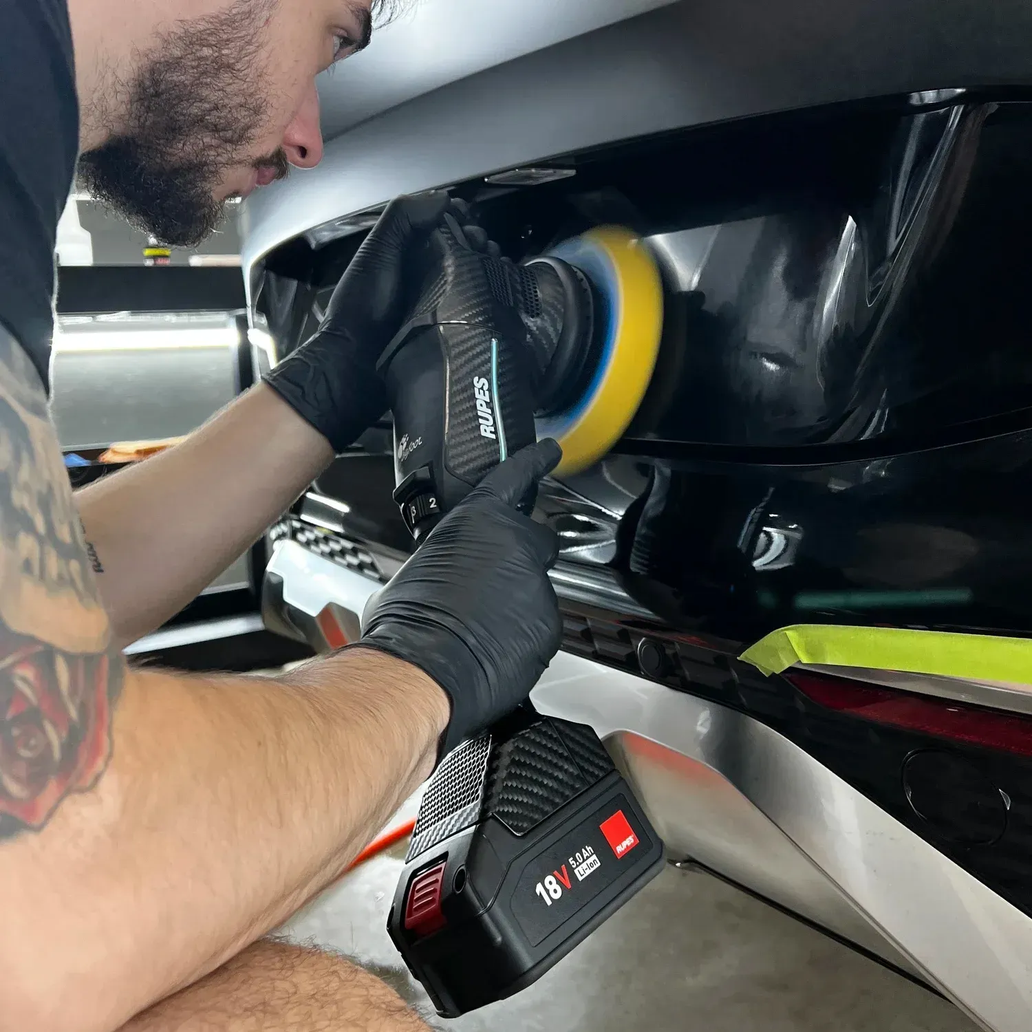 Person polishing a black car panel with a yellow buffer in a garage
