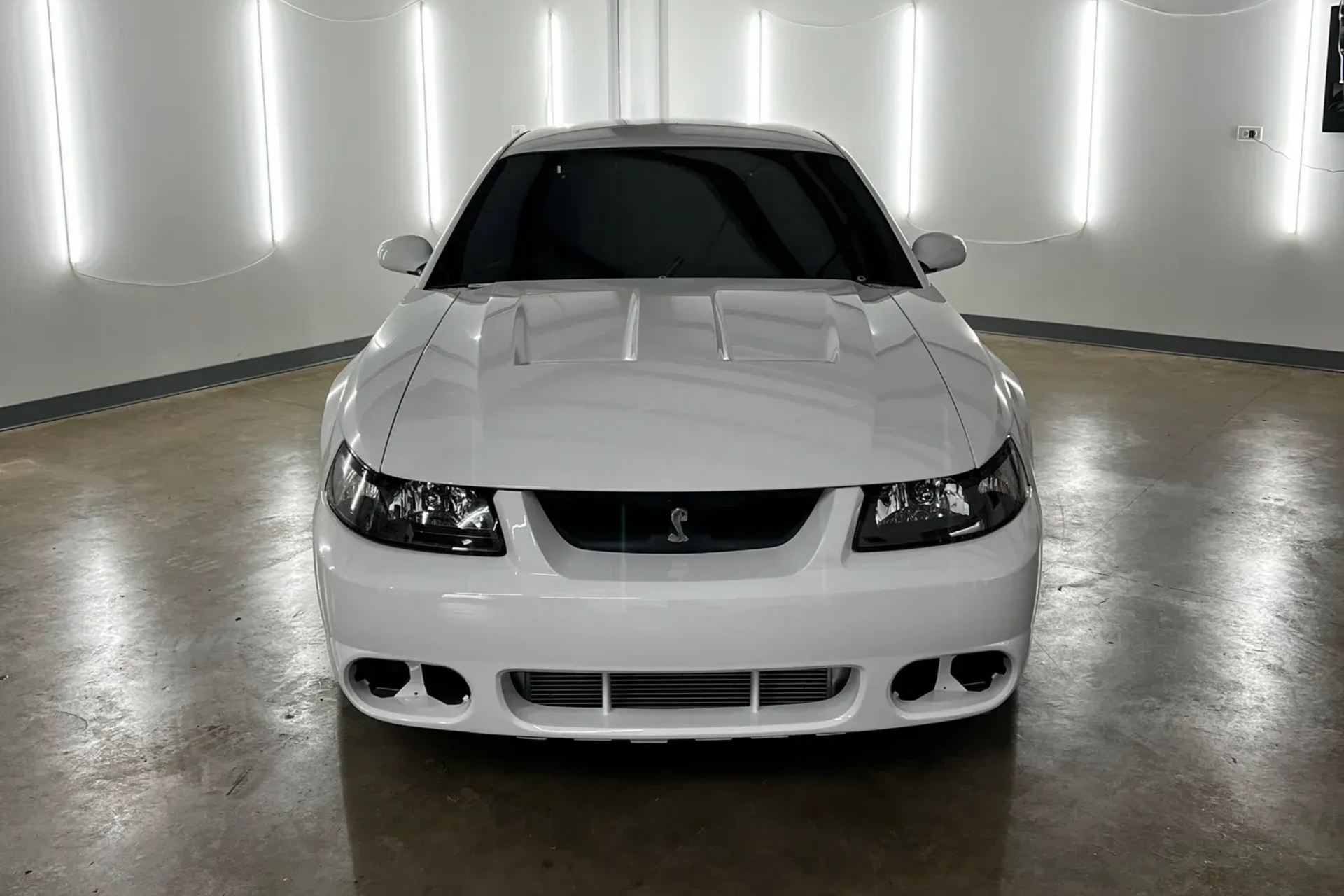 White sports sedan front view in a studio, with black hood and headlights on, against lit wall panels.