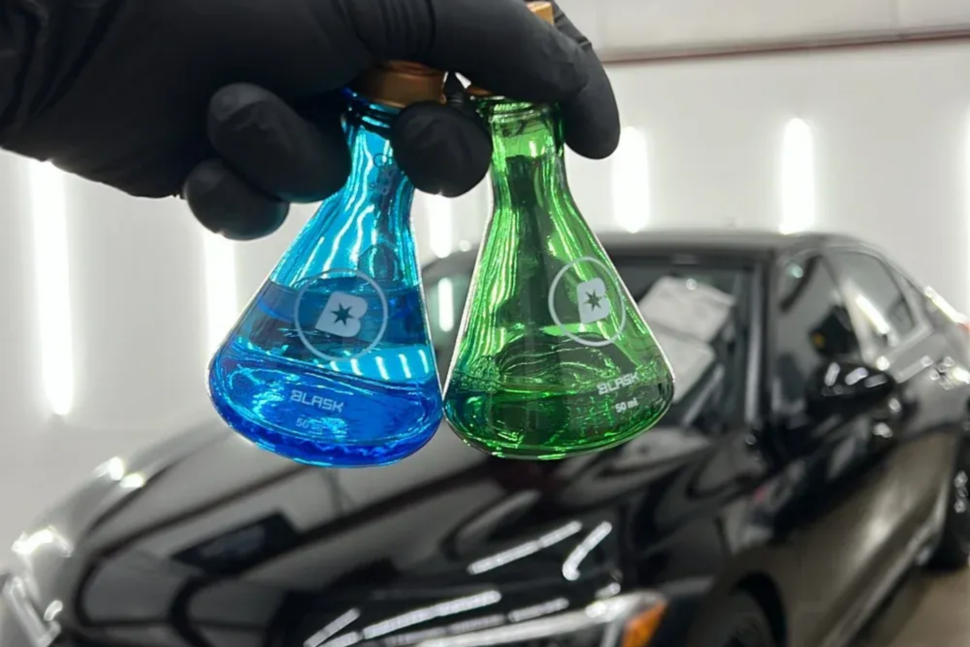 Gloved hand holding blue and green car-cleaning solution bottles in front of a black car