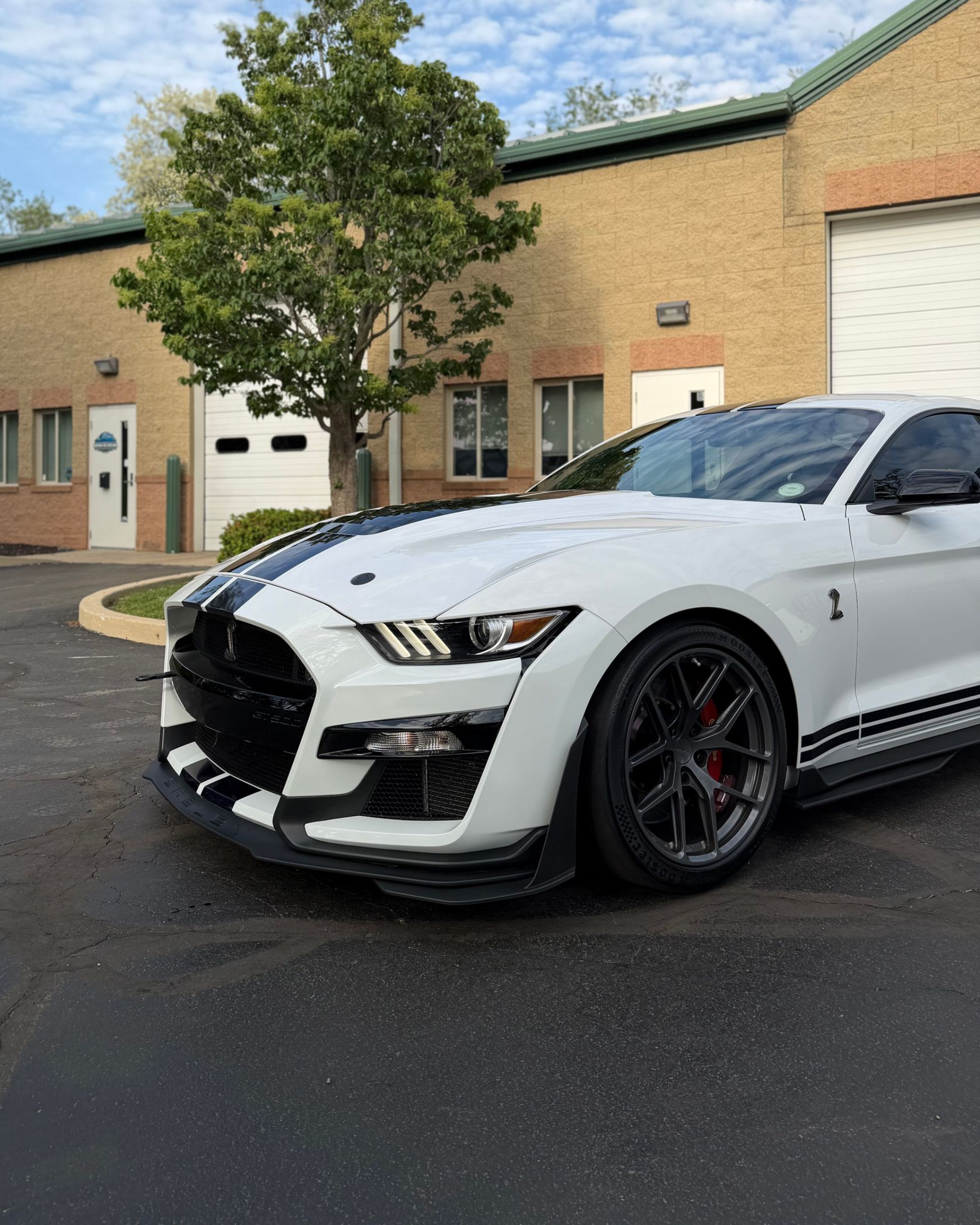 White sports car parked outside a brick building, front angled view on wet pavement.