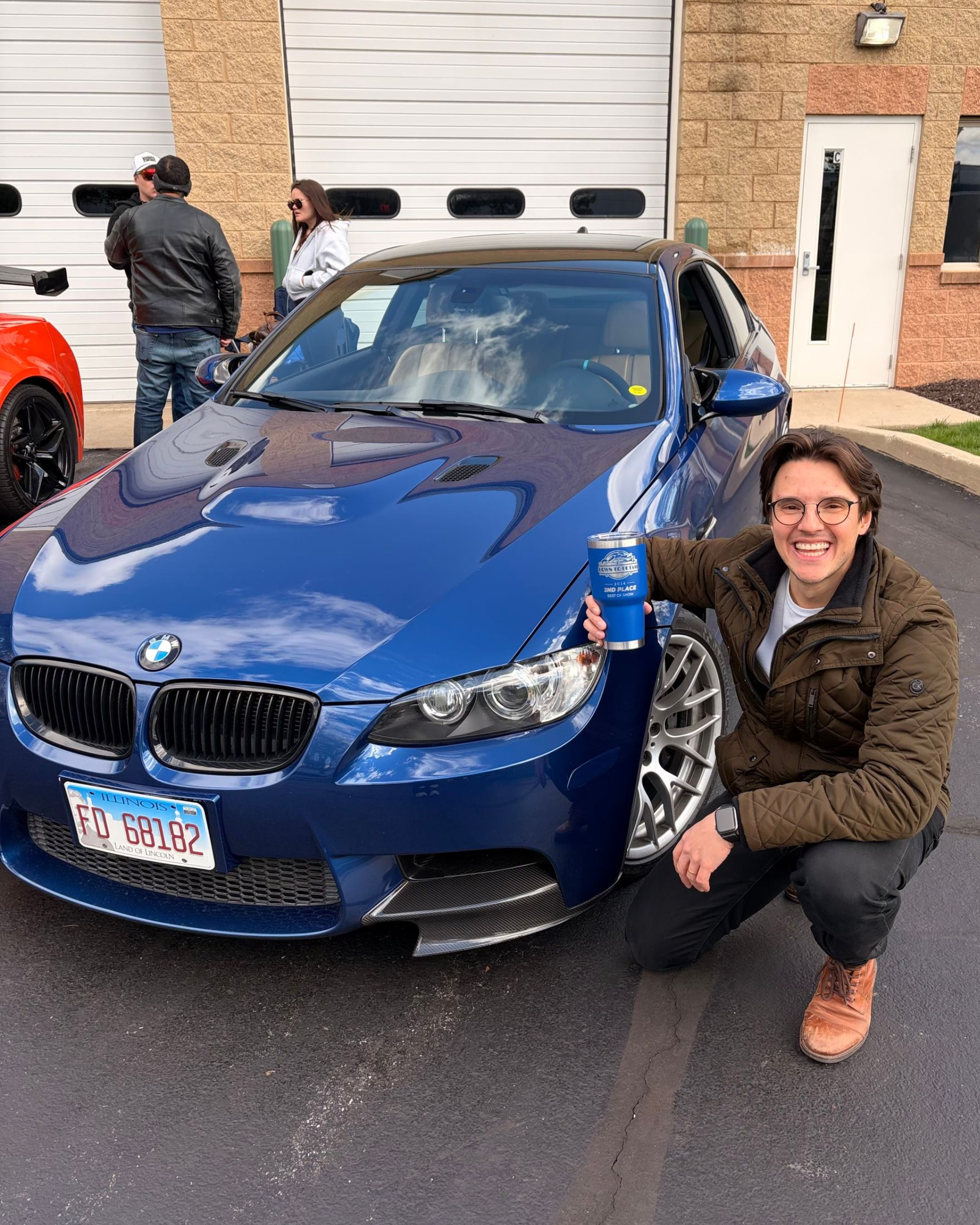 Woman crouching beside a blue BMW sedan in a driveway, smiling at the camera.
