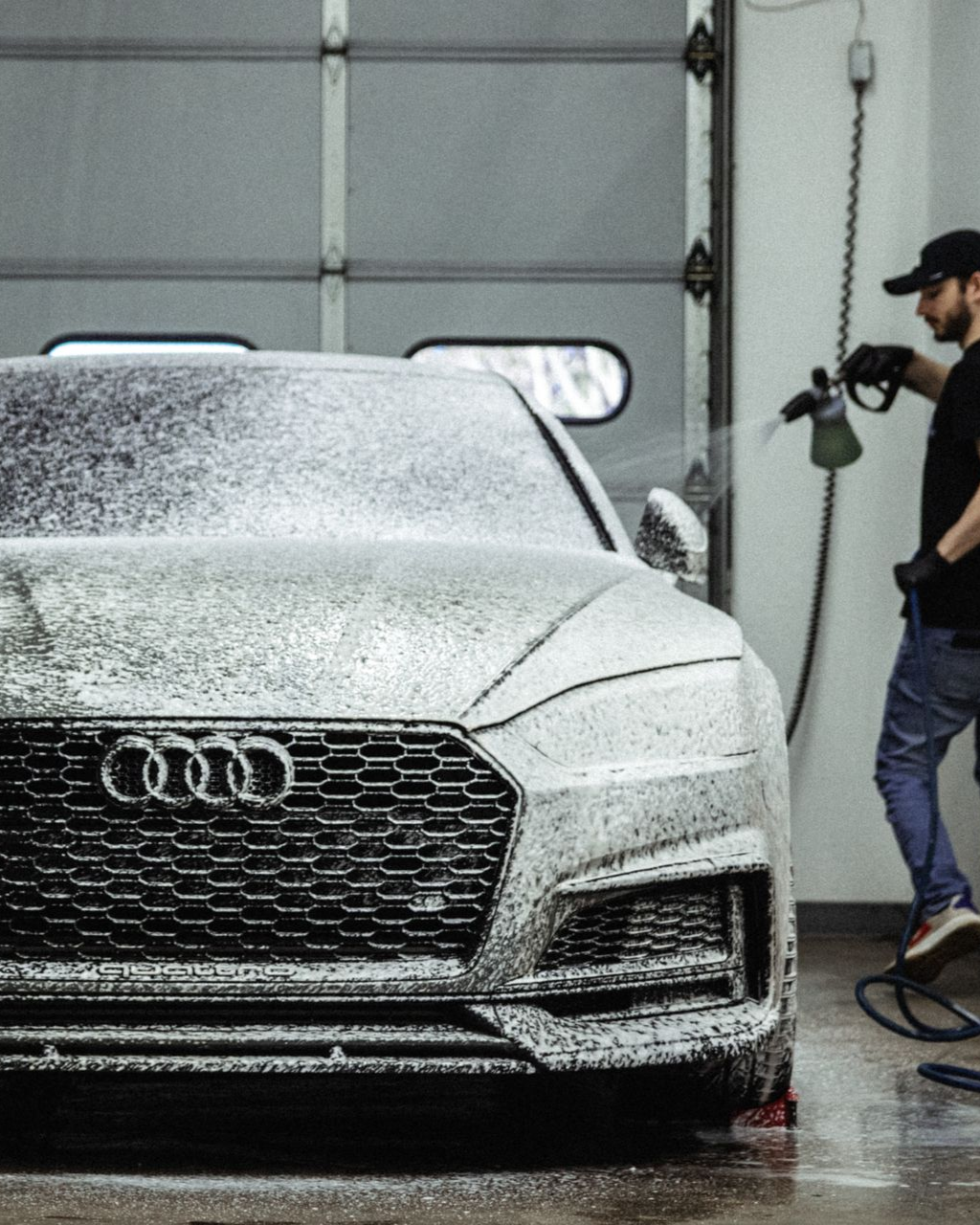 White Audi SUV being sprayed with foam in a car wash while a worker stands nearby