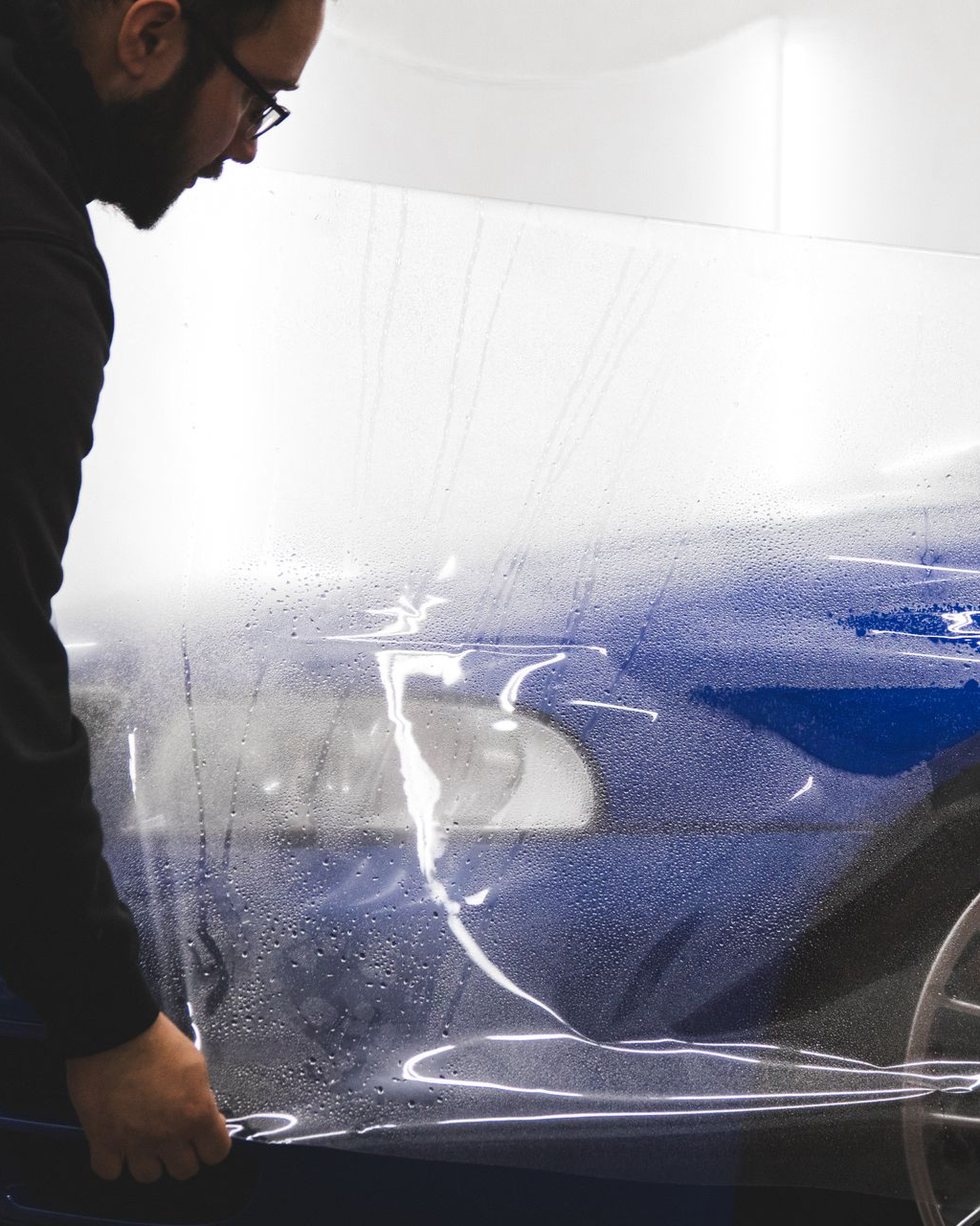 Person holding clear plastic wrap over a blue car in a workshop