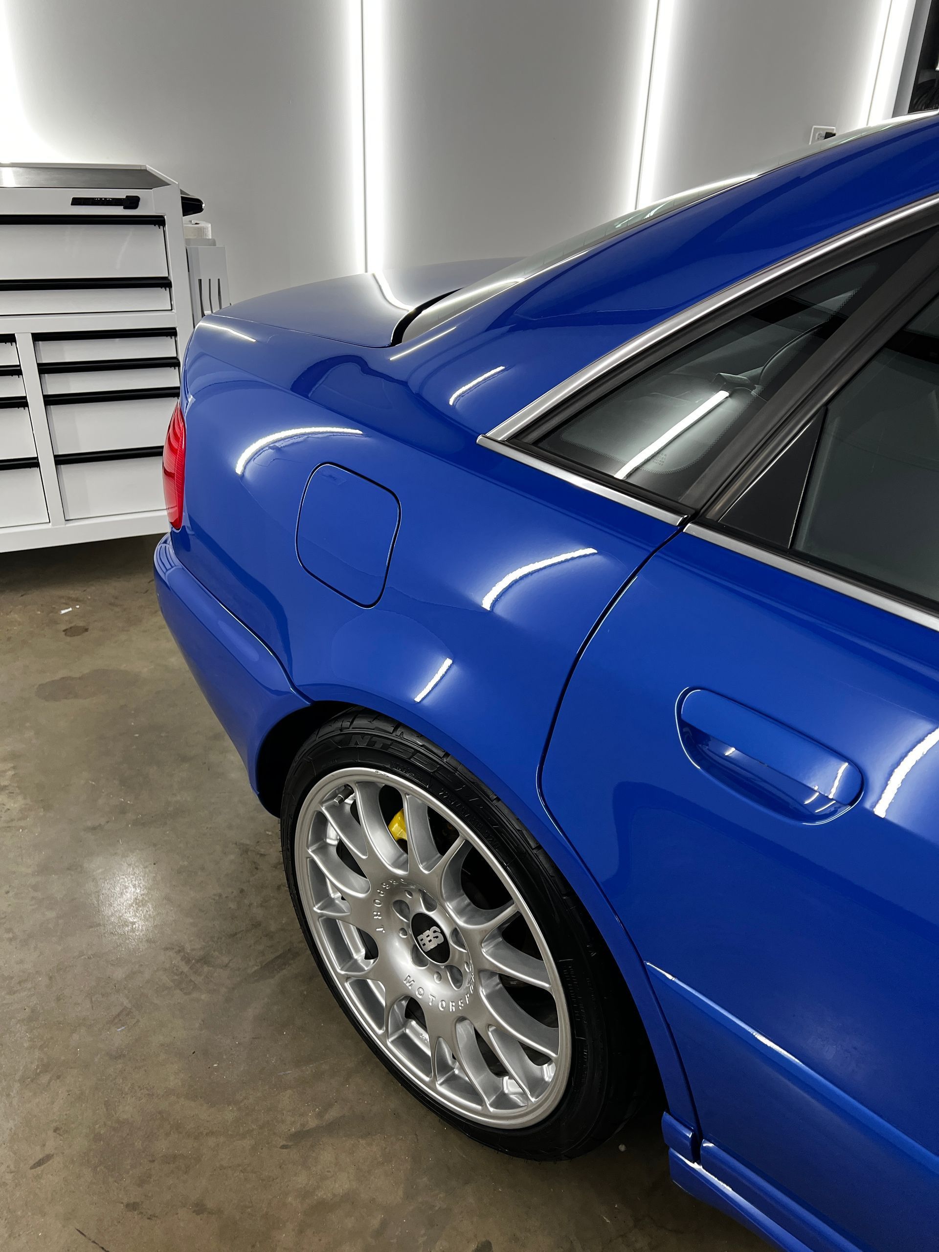 Blue sports car parked in a garage, showing the rear side and silver wheels.