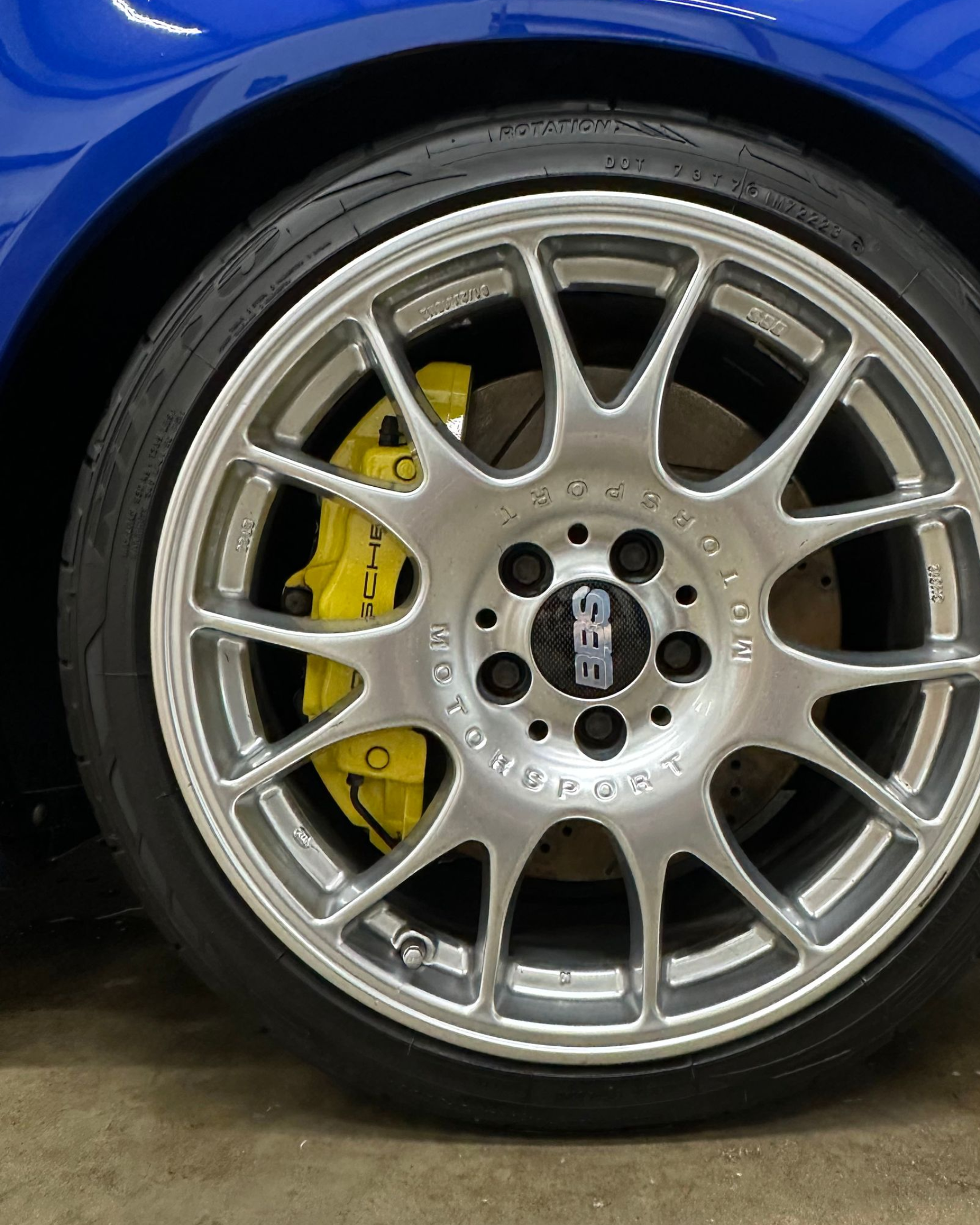 Close-up of a blue car’s silver alloy wheel with yellow brake caliper on a concrete floor