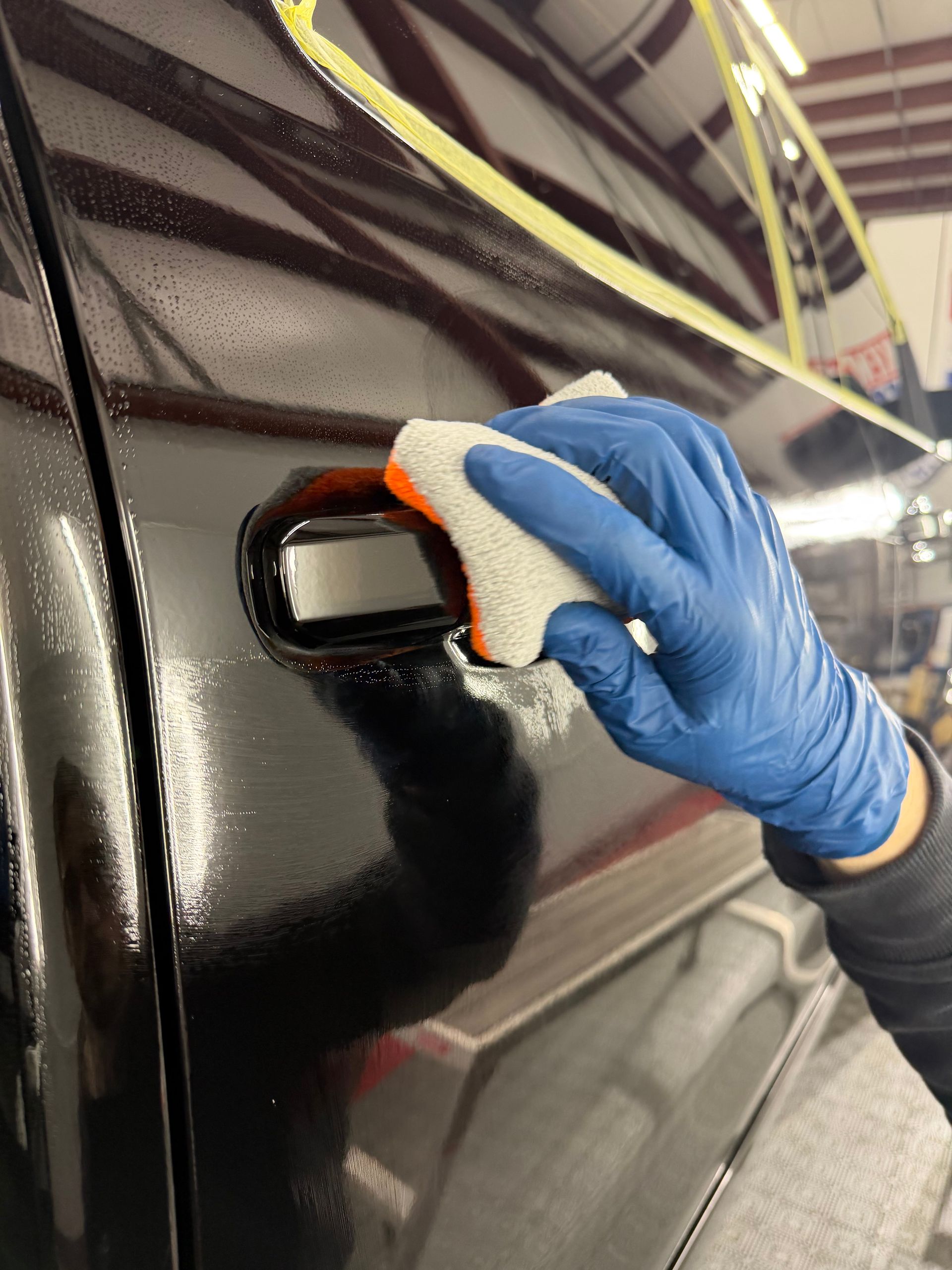 Gloved hand polishing a black car door handle with a cloth in a workshop.