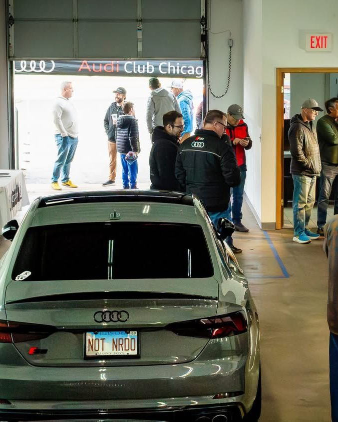 Cars and people gathered in a bright indoor showroom near an Audi Club Chicago sign.