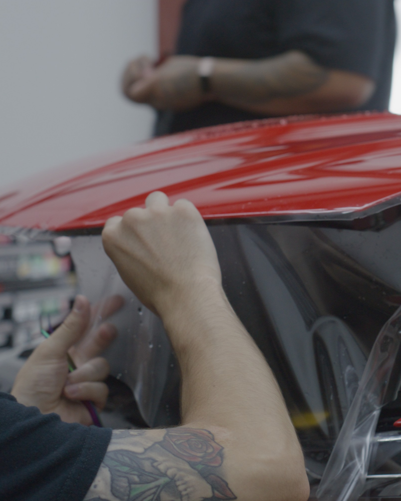 Hands smoothing clear film onto a red car hood at an auto detailing shop