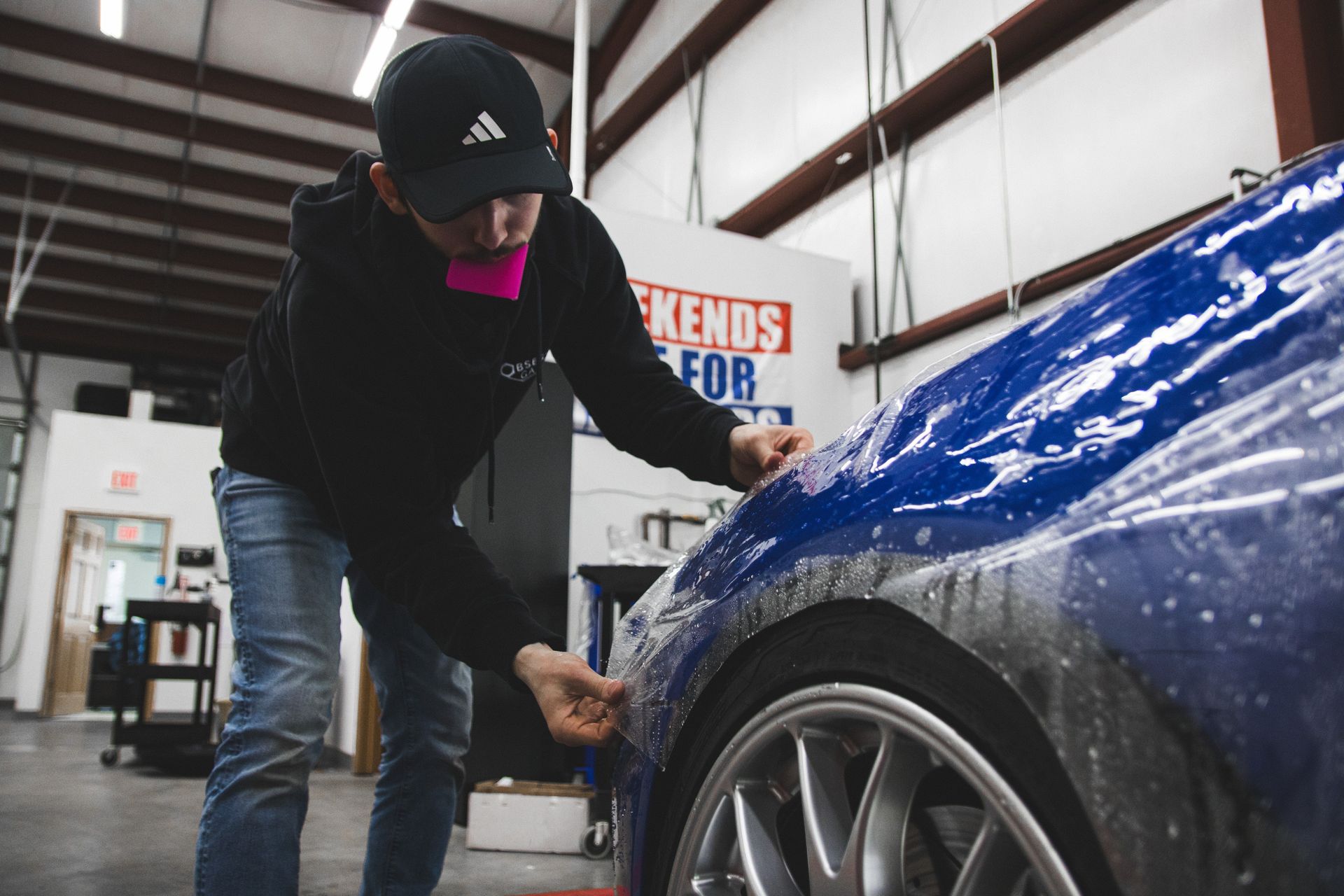 Person detailing a blue car in a garage, polishing the rear quarter panel near the wheel.