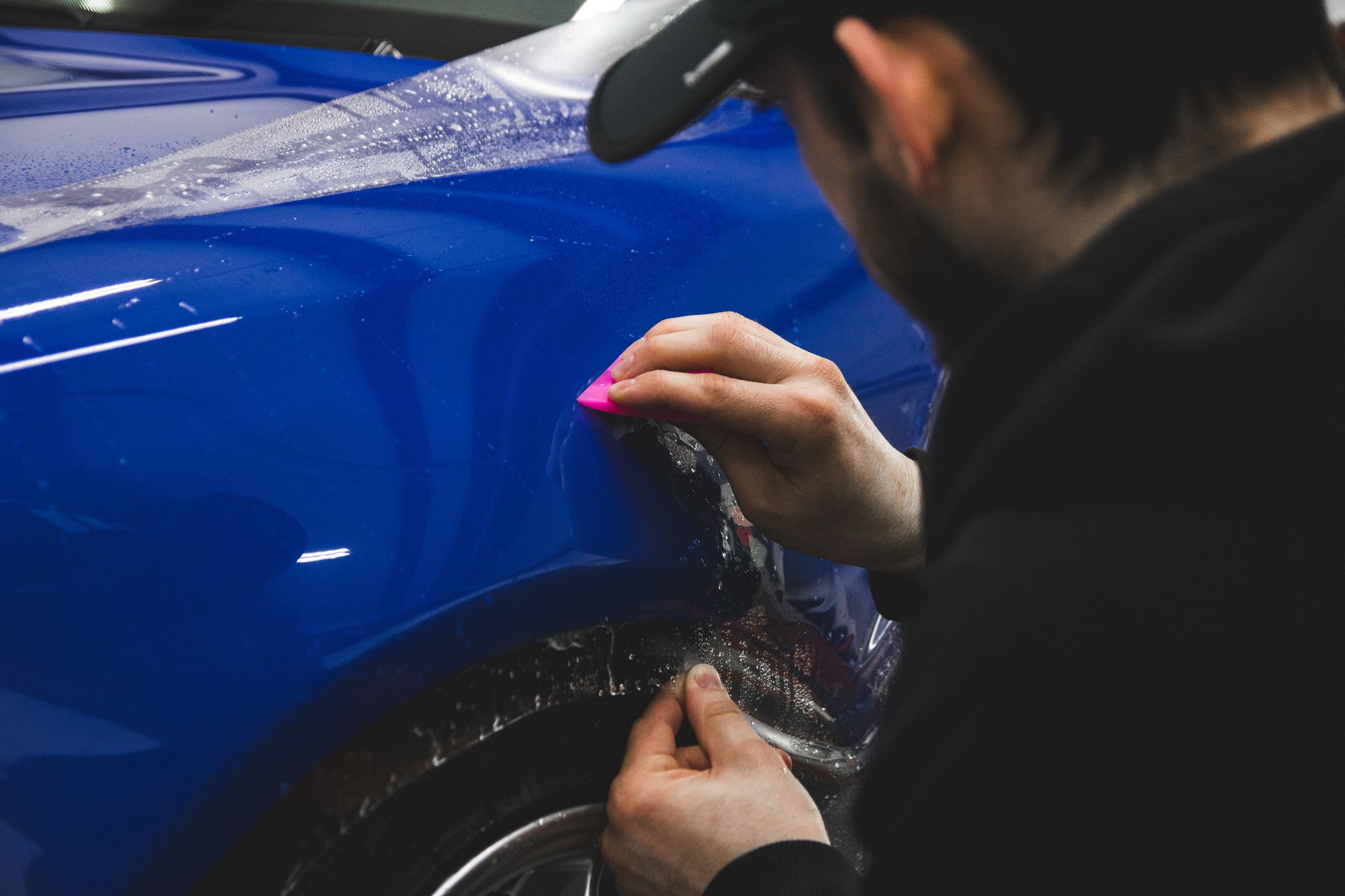 Person applying clear protective film to a blue car panel with a pink squeegee